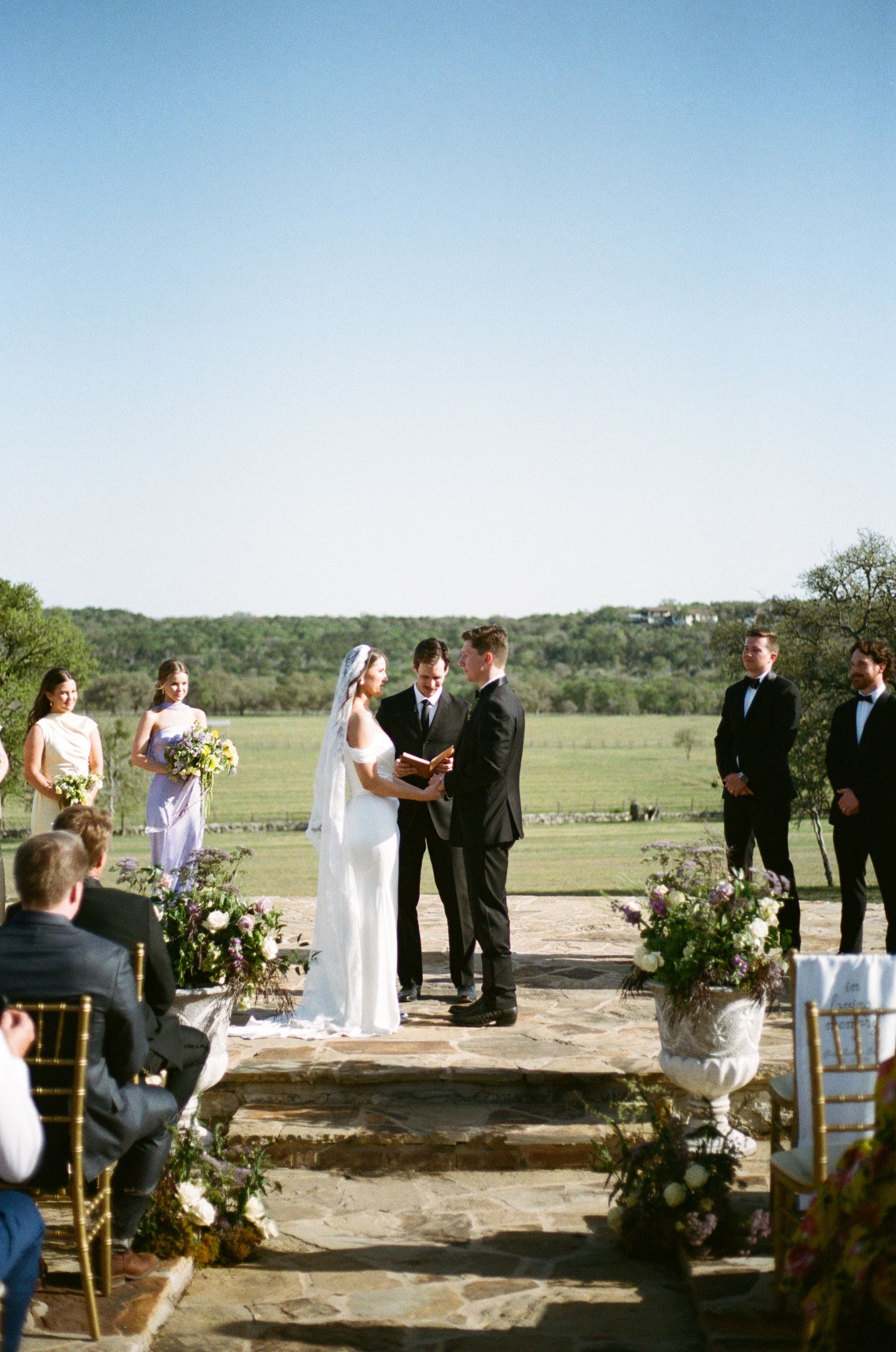 Outdoor wedding ceremony on a stone platform with floral decorations and rolling hills in the background.