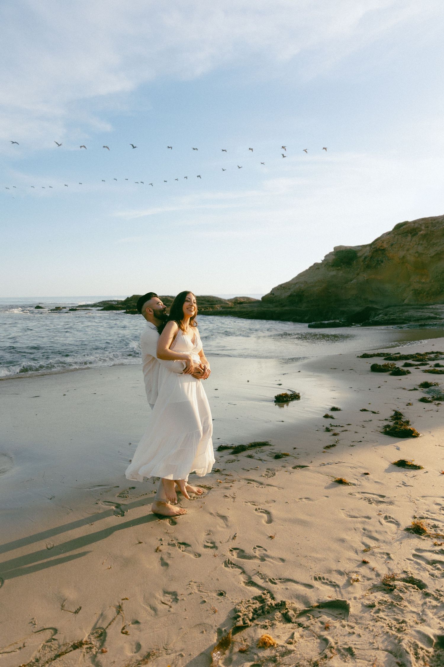 Two people in white dresses walk along a scenic beach at sunset with birds flying overhead in the distance.