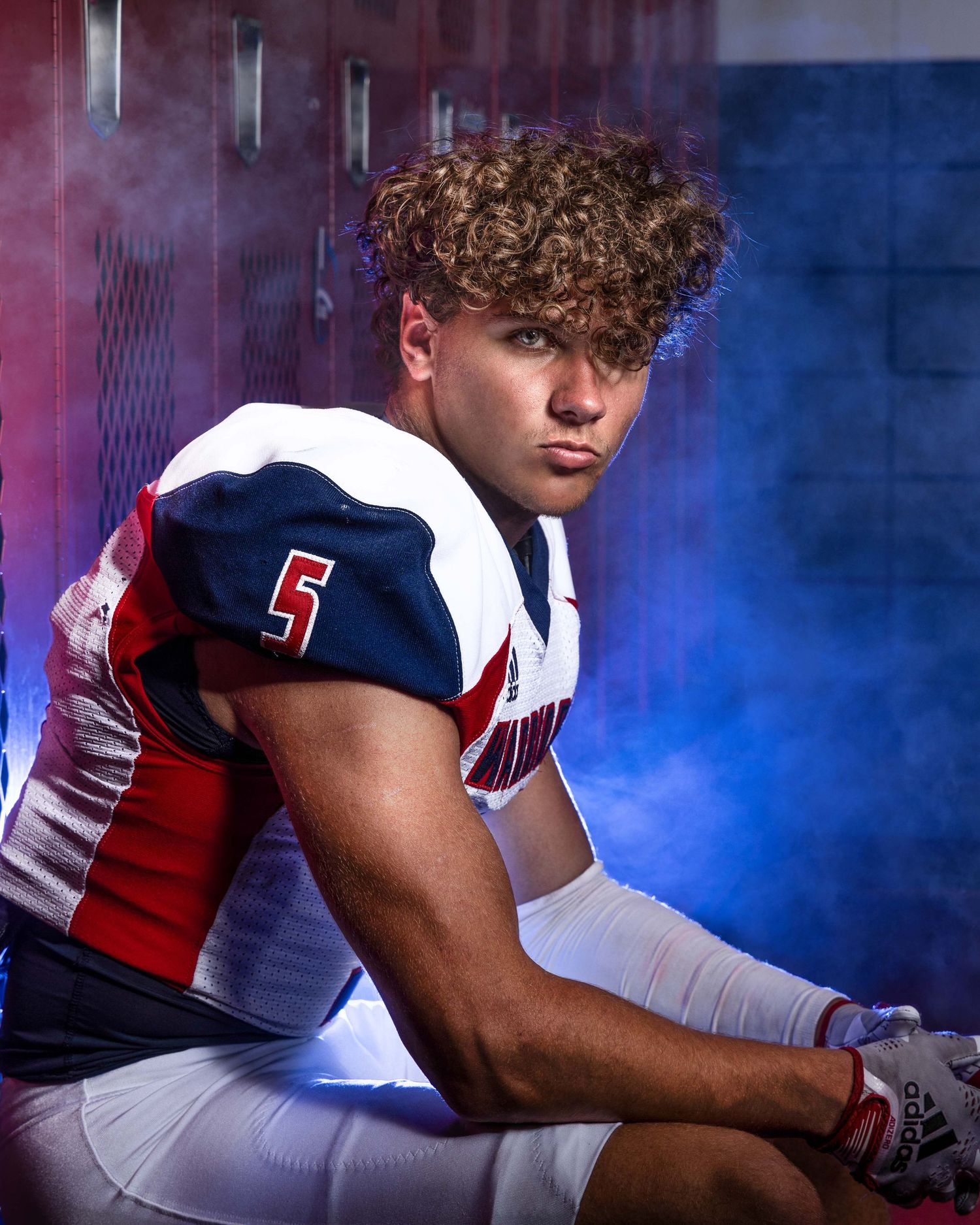 Football player in uniform number 5 sits contemplatively by lockers in dramatic lighting.