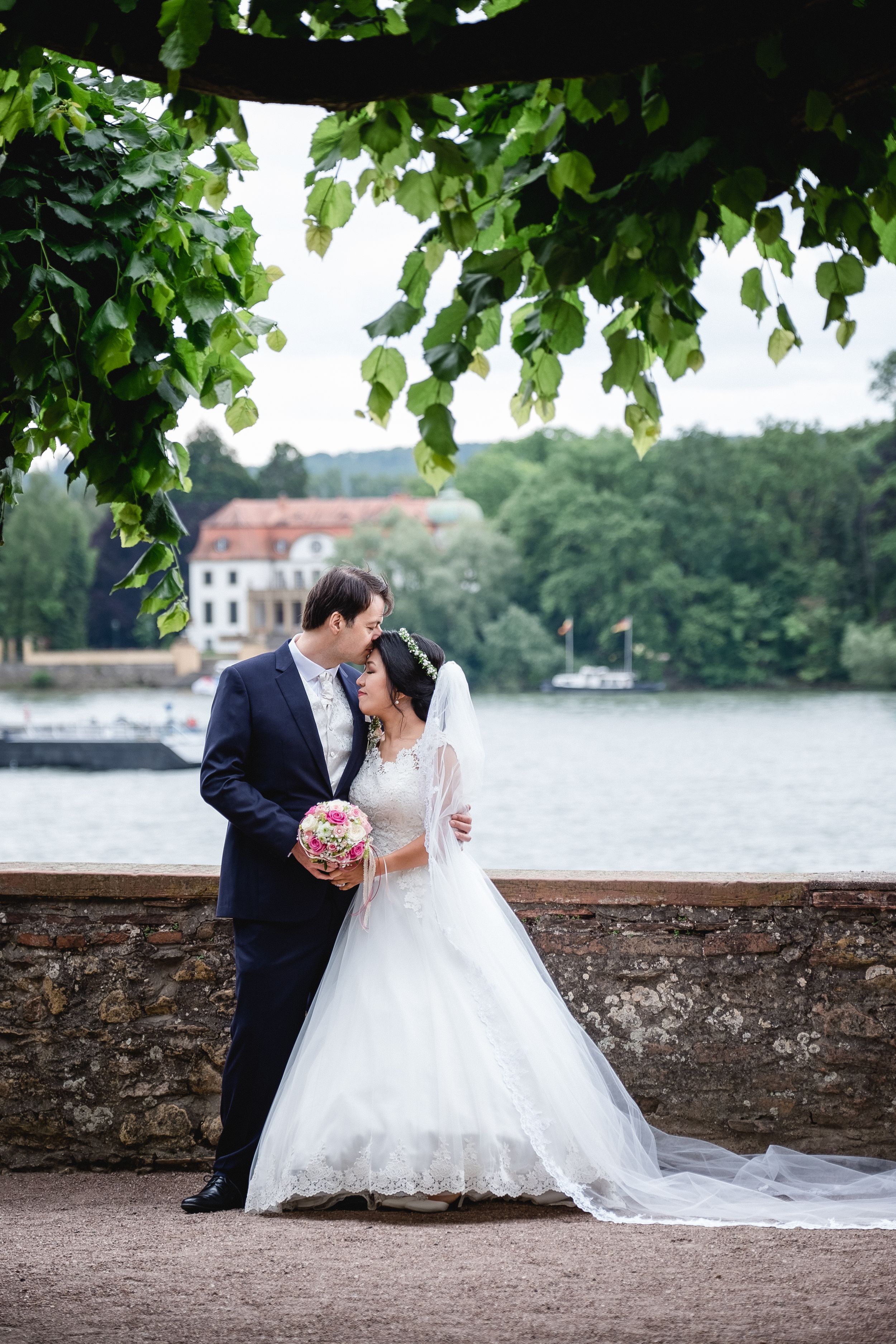 Newlyweds share a romantic moment by a lakeside with green foliage and historic building in background.