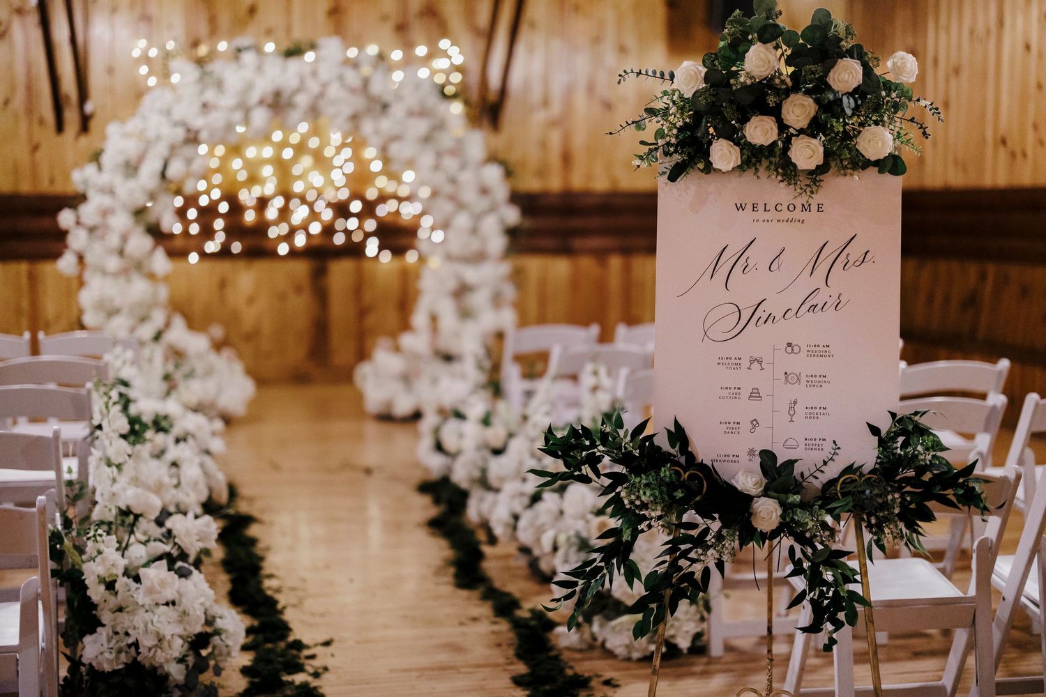 Elegant wedding ceremony setup with white floral arch, twinkle lights, and a blush welcome sign in a rustic wooden venue.