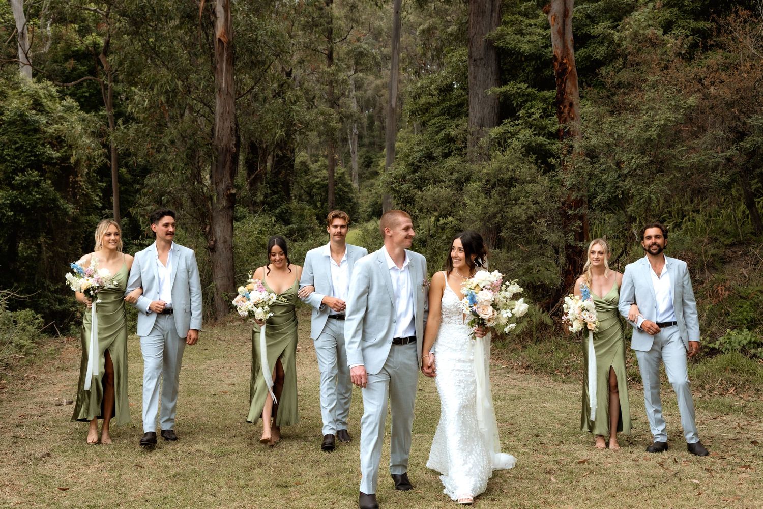Wedding party in sage green and light blue attire walk through a wooded forest setting with white floral bouquets.