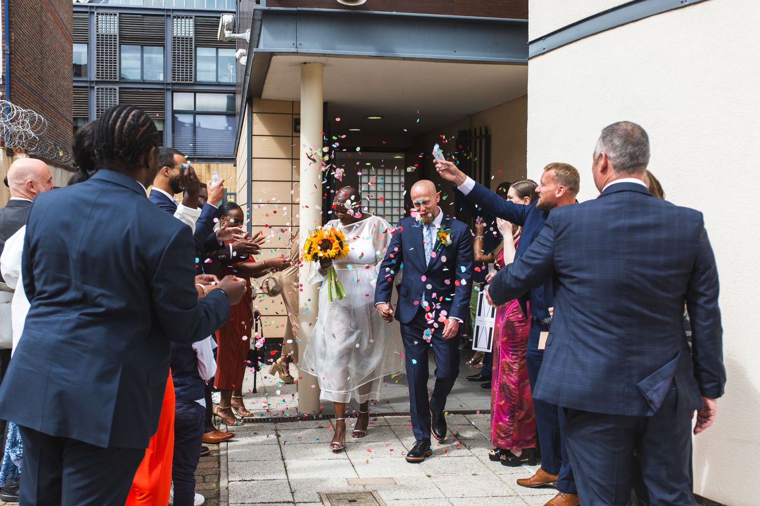 Wedding party celebrating with confetti outside building entrance.
