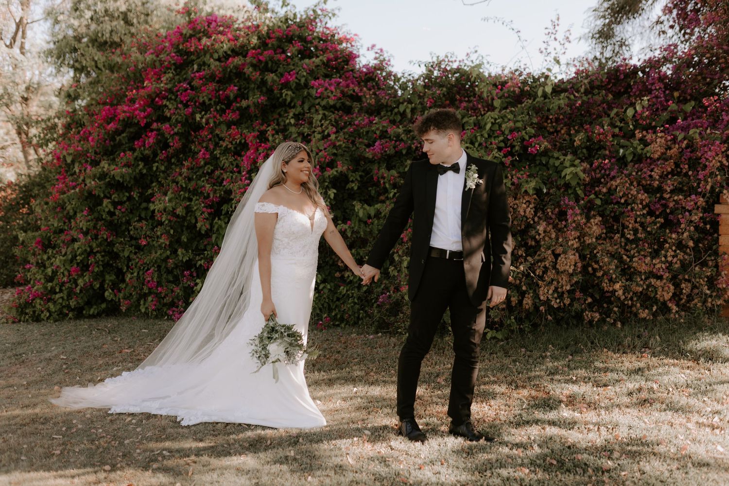 A romantic wedding photo of a couple posing against a flowering bougainvillea backdrop in formal attire.