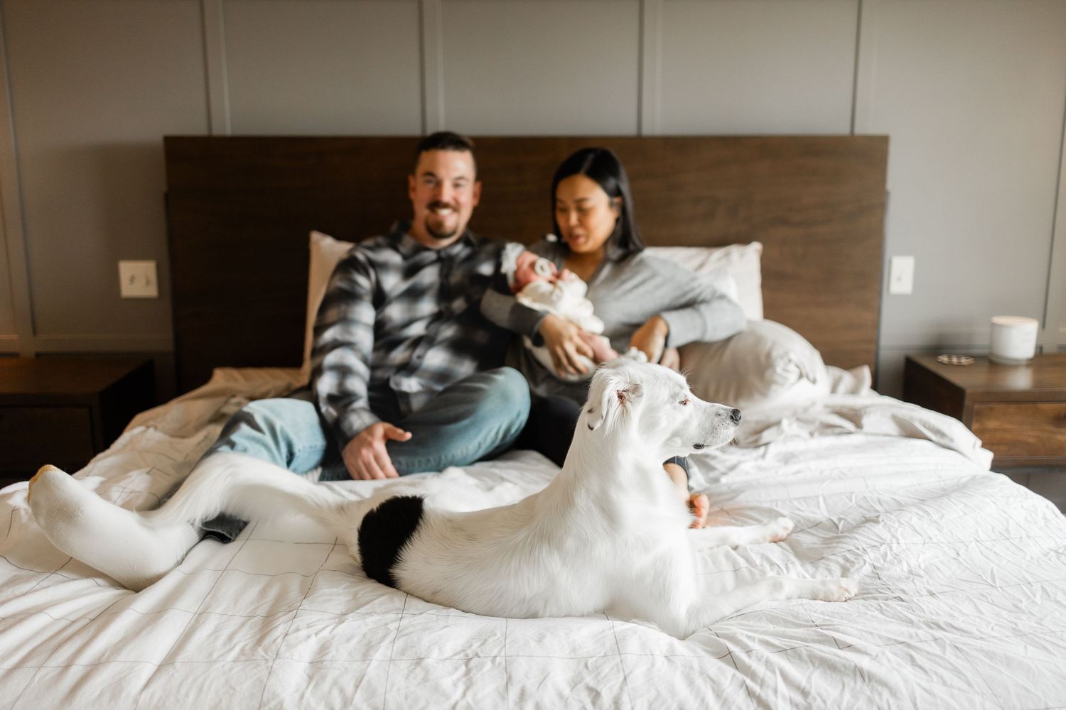A couple relaxes on a bed with their white dog in a cozy bedroom setting.