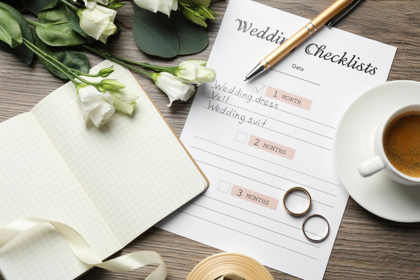 Flat lay of wedding planning items including rings, checklist, flowers and coffee on wooden table.