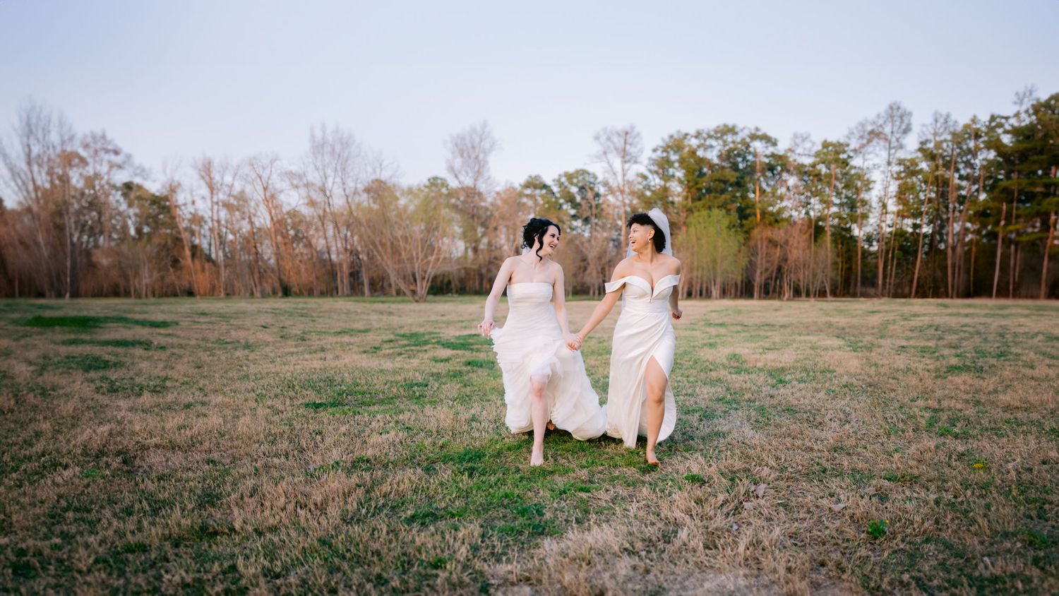 Two people in white dresses run together across a grassy field with trees in the background at sunset.