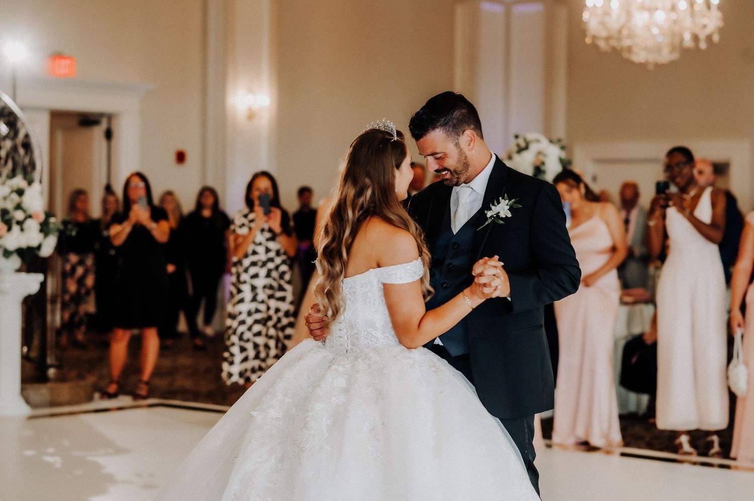 A romantic first dance between newlyweds in an elegant ballroom with a sparkling chandelier and guests watching.