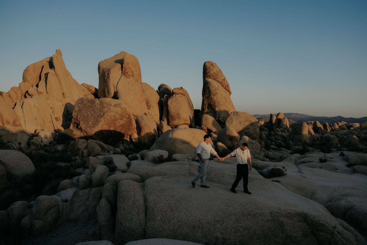 Two figures walk among large rock formations in desert landscape at sunset.
