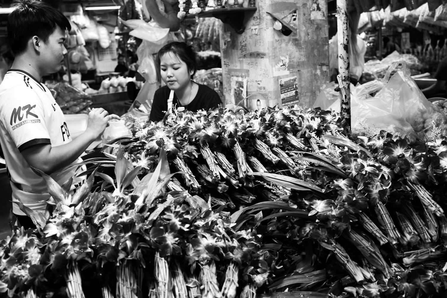 Black and white market scene with vendors selling large bundles of fresh vegetables at an outdoor stall.