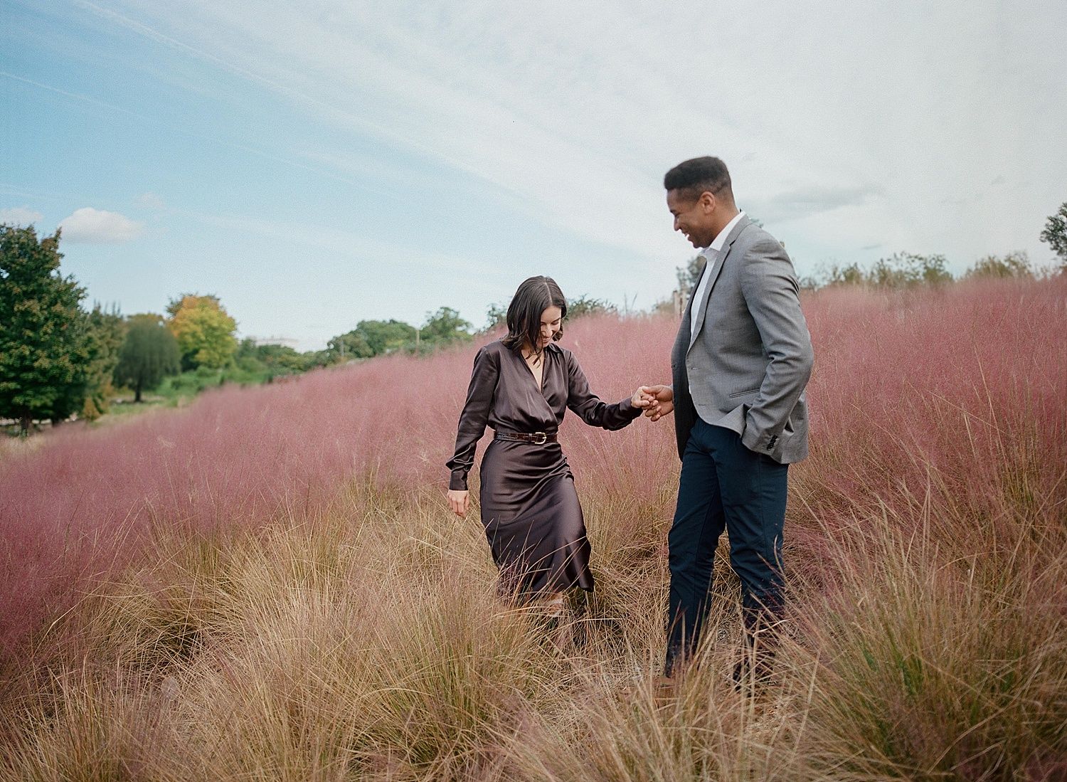 A couple shares a tender moment while standing in a field of pink ornamental grass against a bright blue sky.