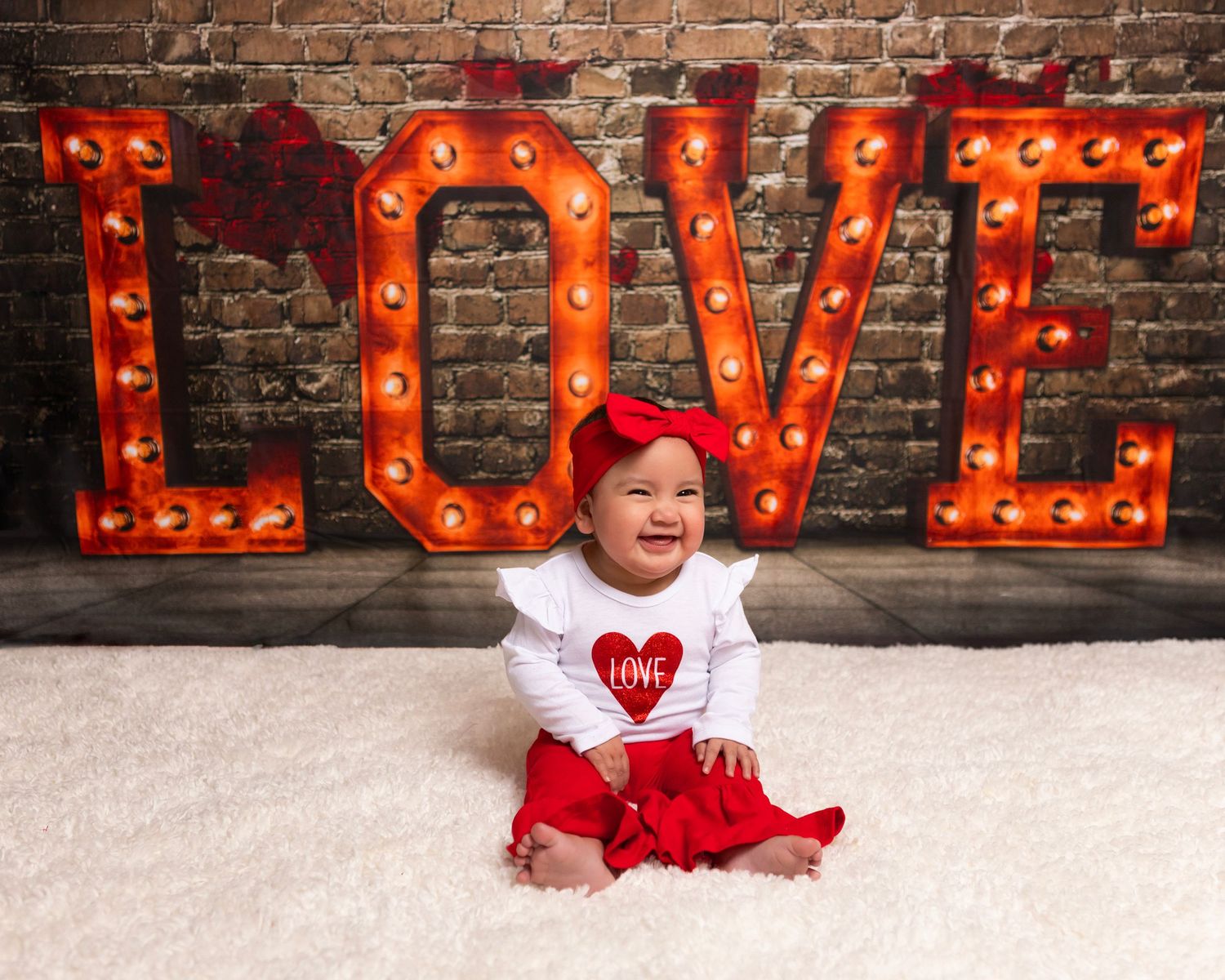 Valentine's photo setup with illuminated LOVE marquee letters against brick wall and white floor.