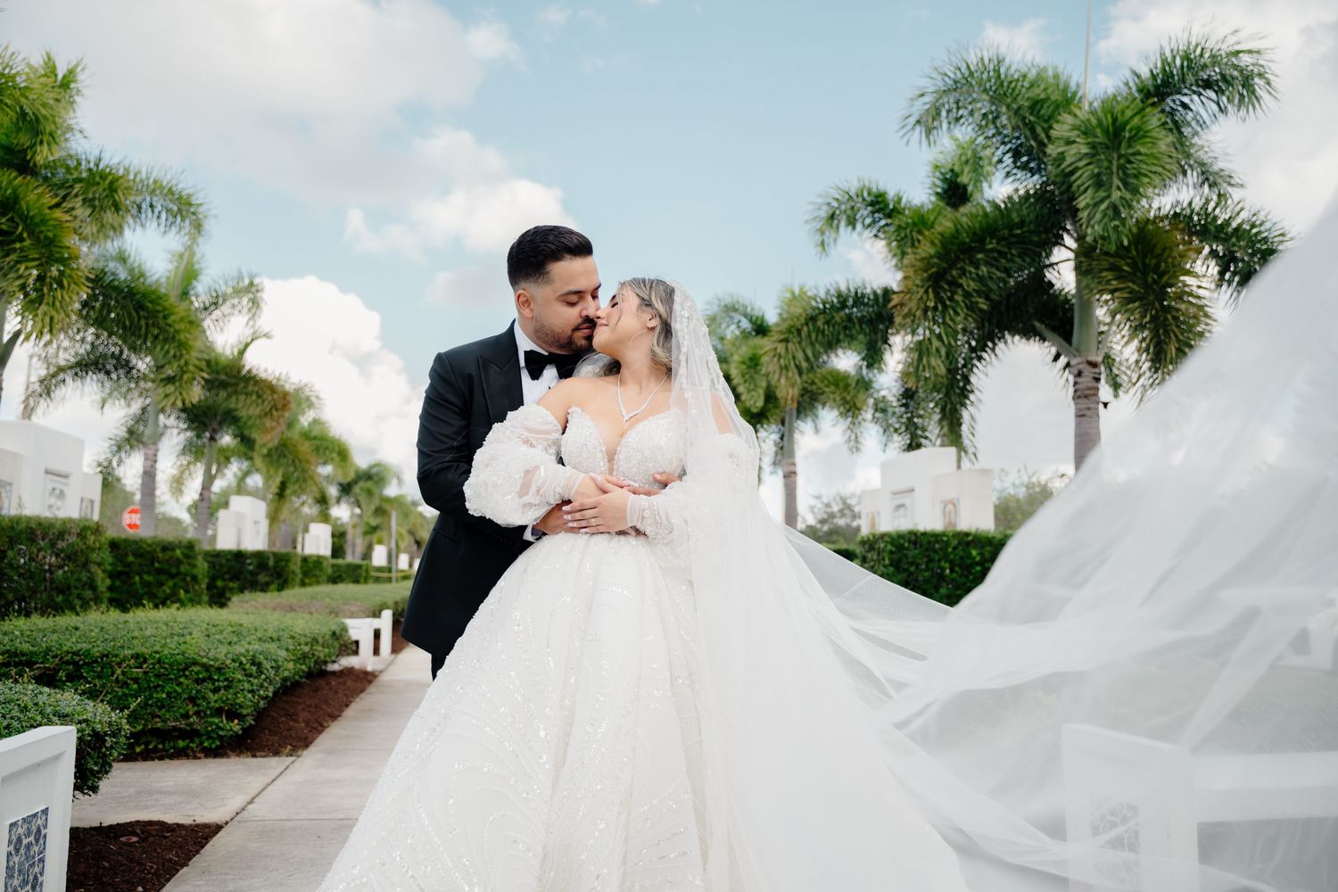 Couple in formal wedding attire embracing outdoors with flowing veil and tropical palm trees in the background.