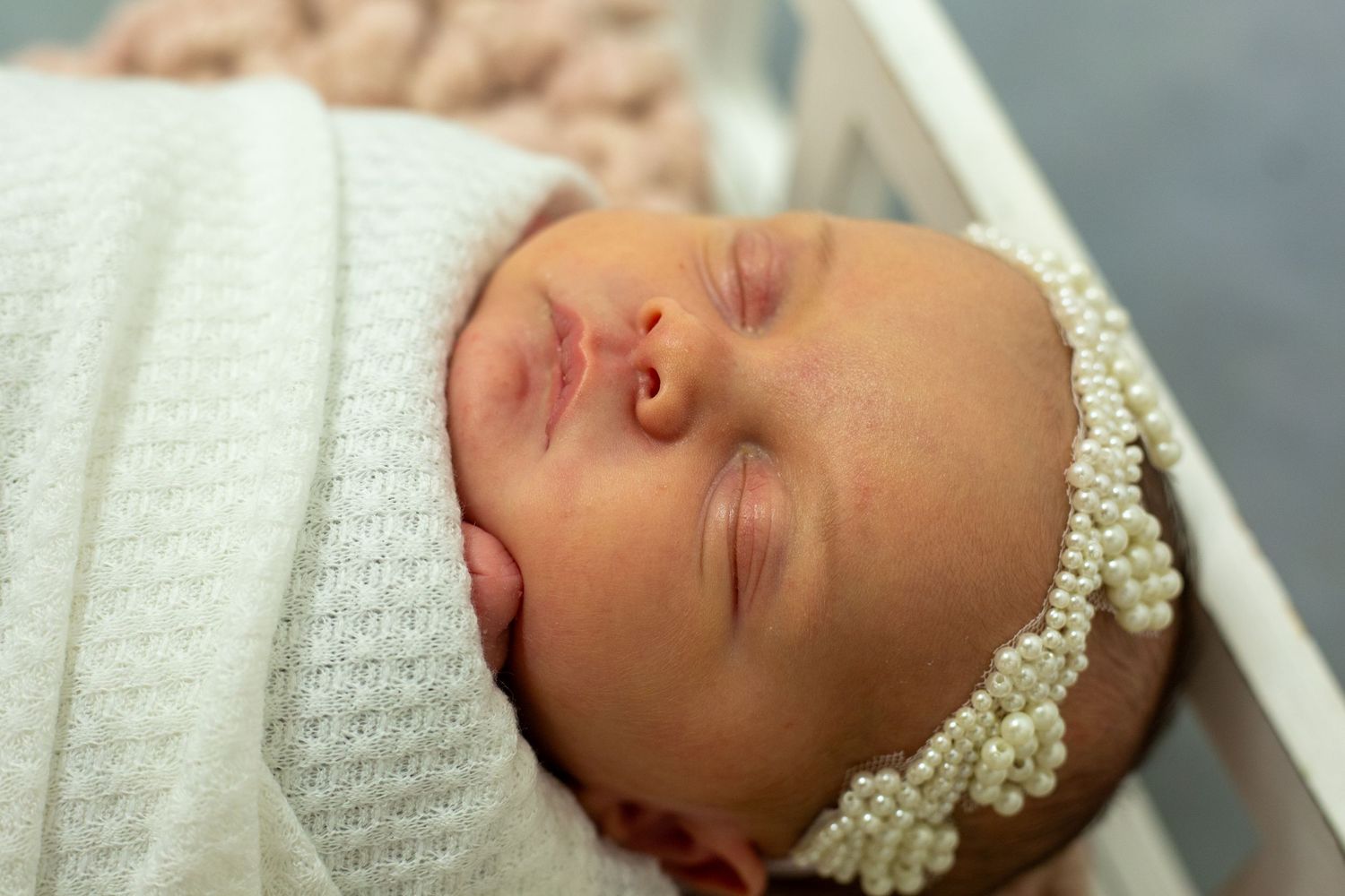 Peaceful sleeping newborn baby wearing a delicate pearl headband and wrapped in a white textured blanket.