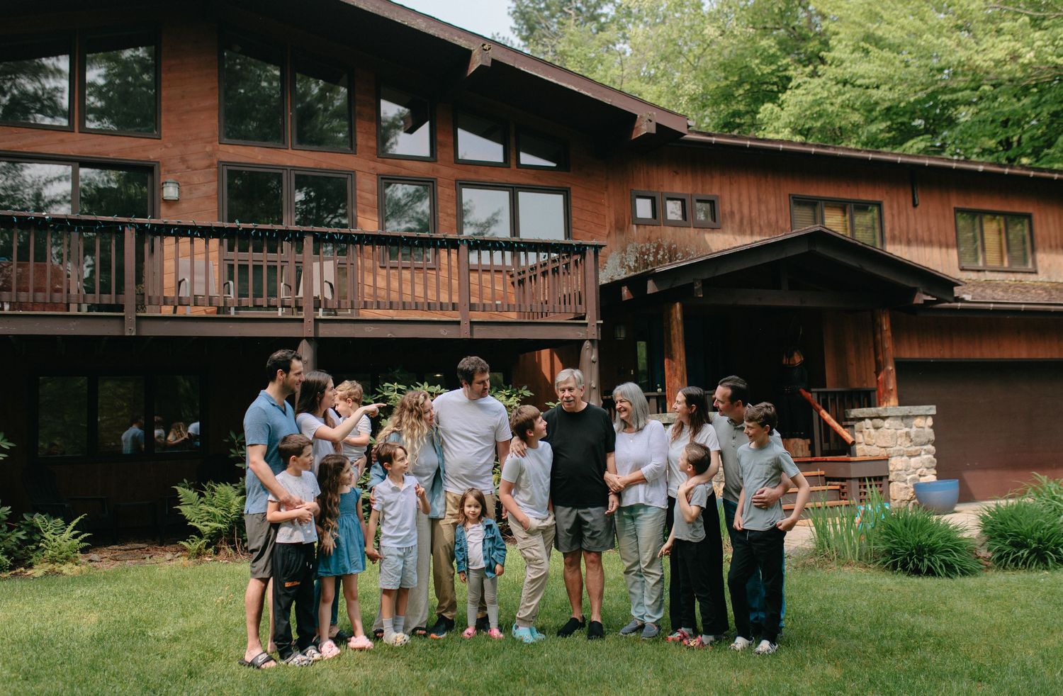 A large rustic wooden house with a deck surrounded by trees serves as backdrop for a casual outdoor gathering.