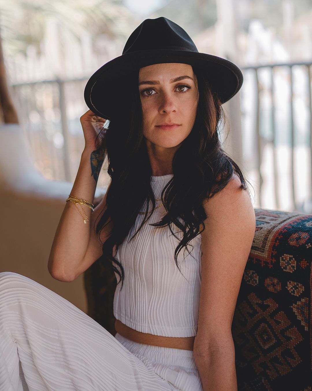 A person in a black wide-brim hat and white crop top sits on a patterned couch with long dark hair.