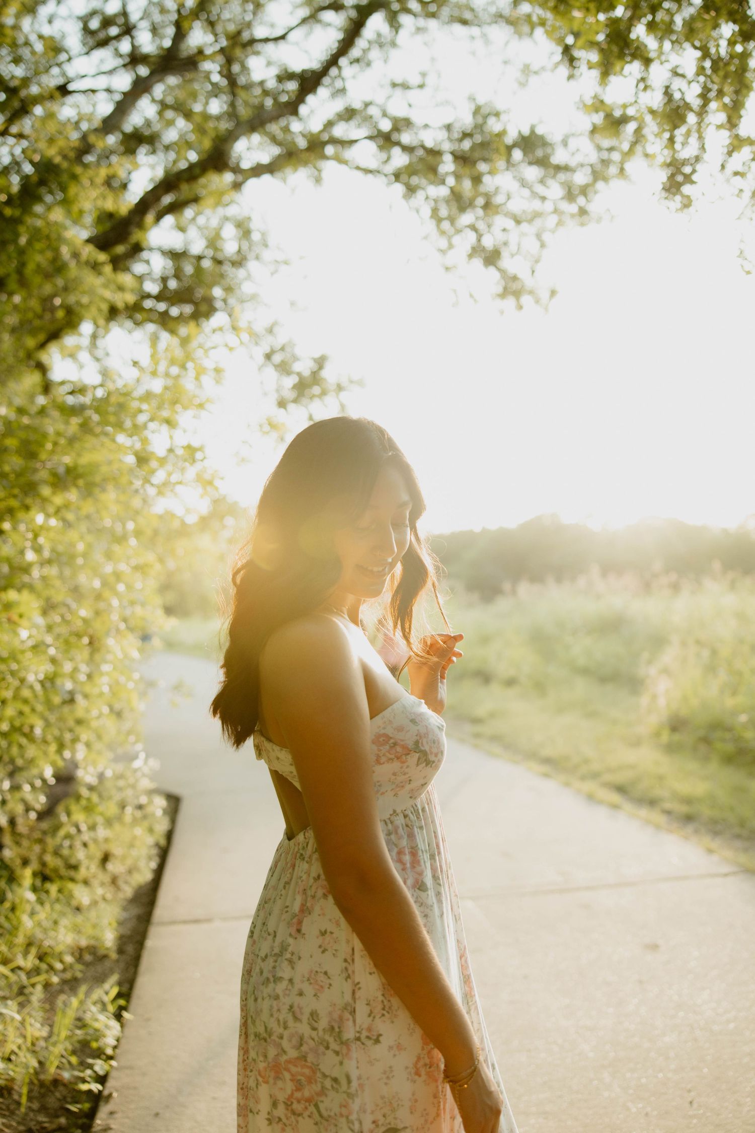 A person in a floral dress stands on a sunlit pathway under trees with golden light streaming through the branches.
