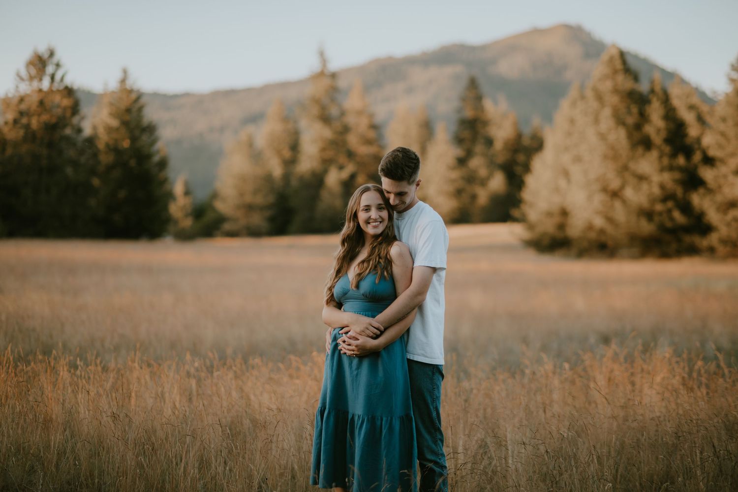 A couple embraces in a golden field at sunset with mountains and evergreen trees in the backdrop.