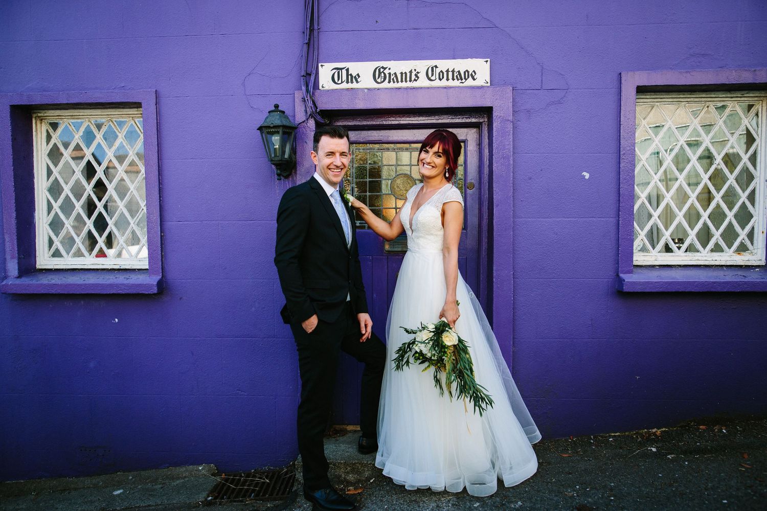 Newlyweds pose outside purple painted quaint storefront with white latticed windows and rustic charm.