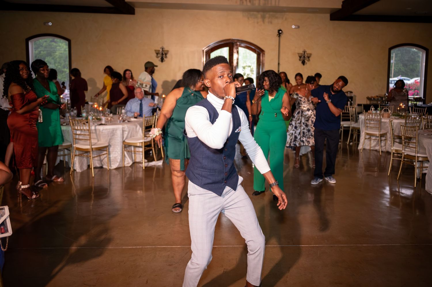 Guest in navy vest and white pants dances energetically on wooden dance floor at reception venue with green decor.