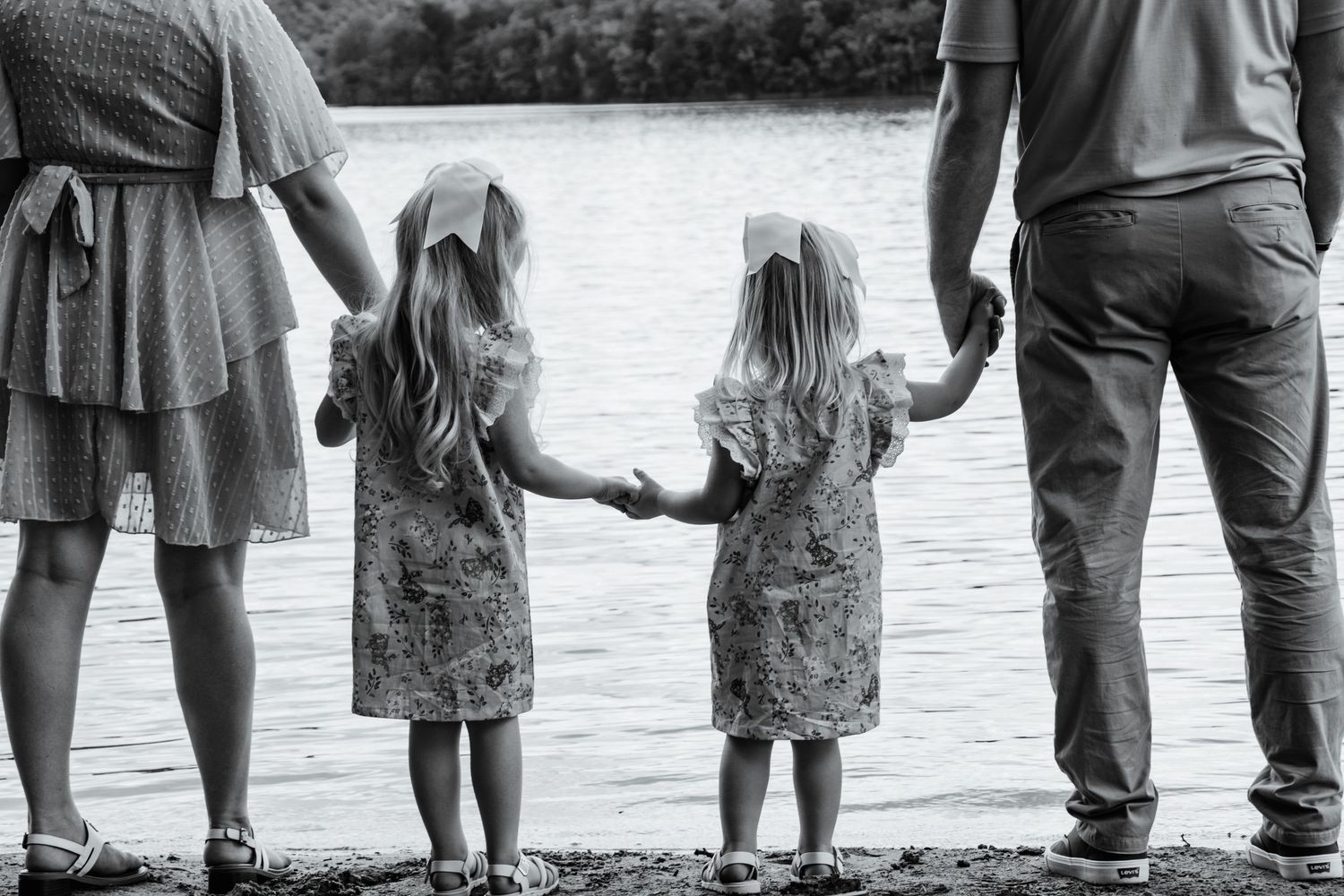 Black & white photo of two little girls holding hands between their parents while standing at the edge of a lake