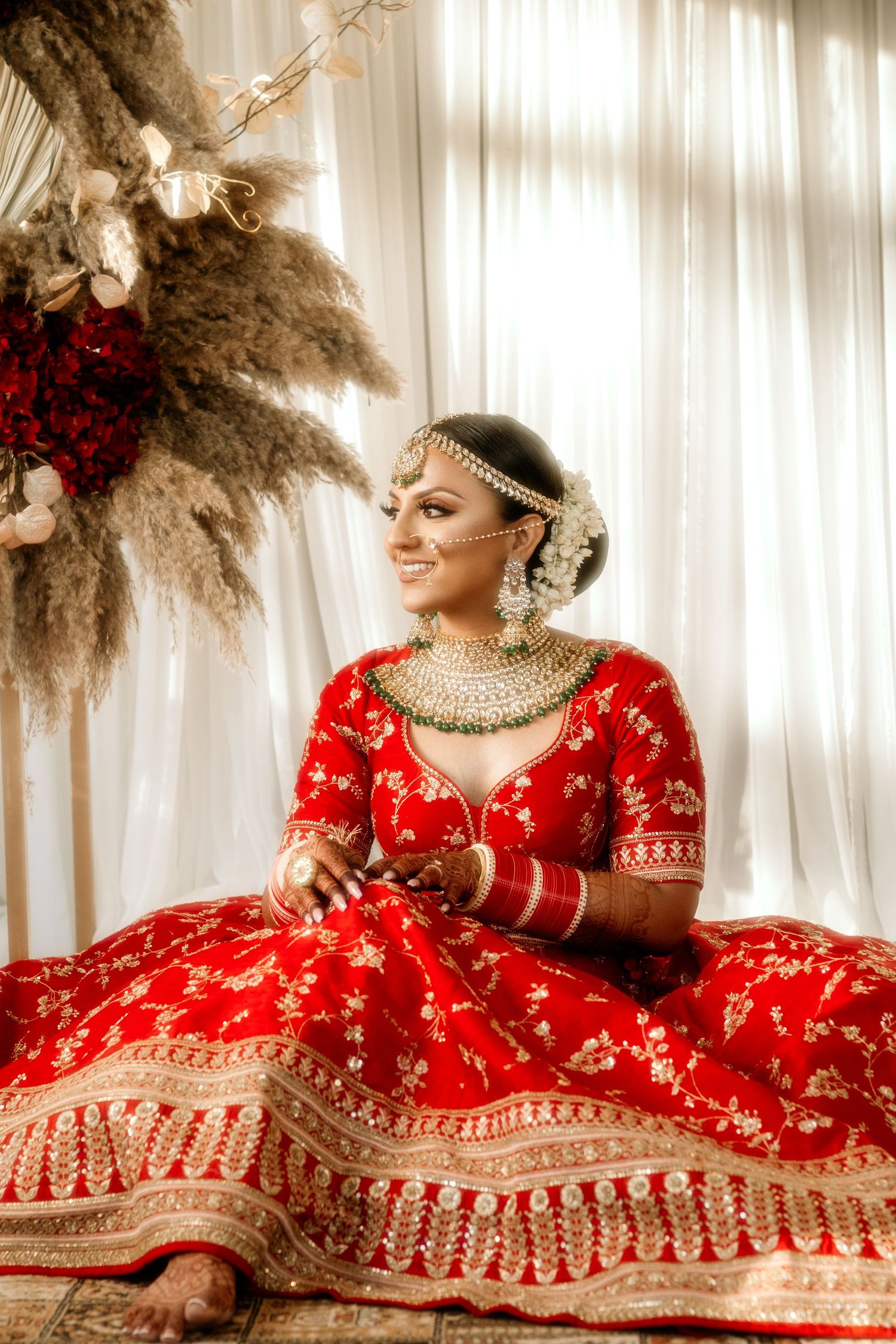 A bride in a traditional red and gold lehenga sits elegantly with pampas grass decor in the background.
