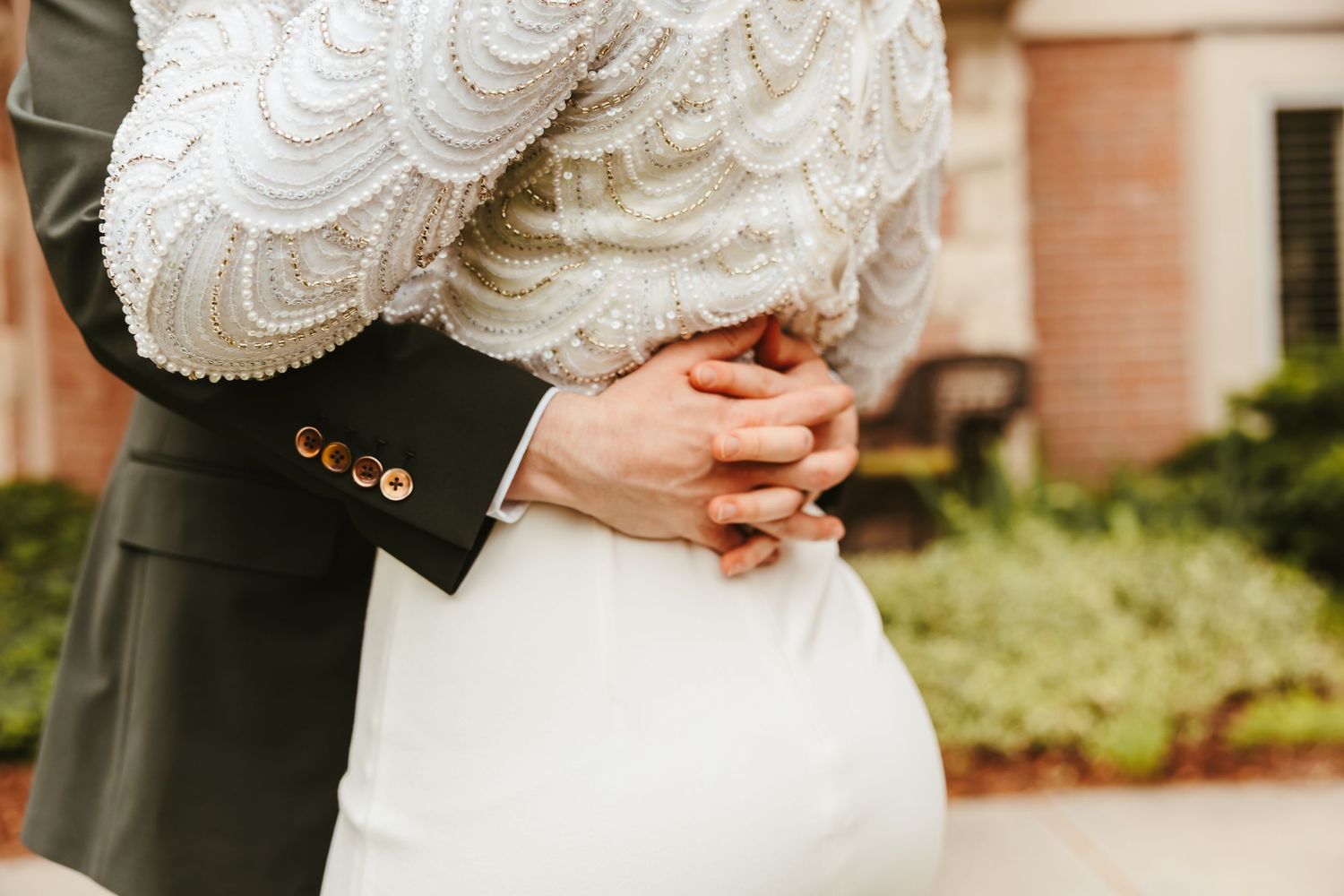 A close-up wedding photo showing clasped hands against a white lace dress and dark suit jacket.