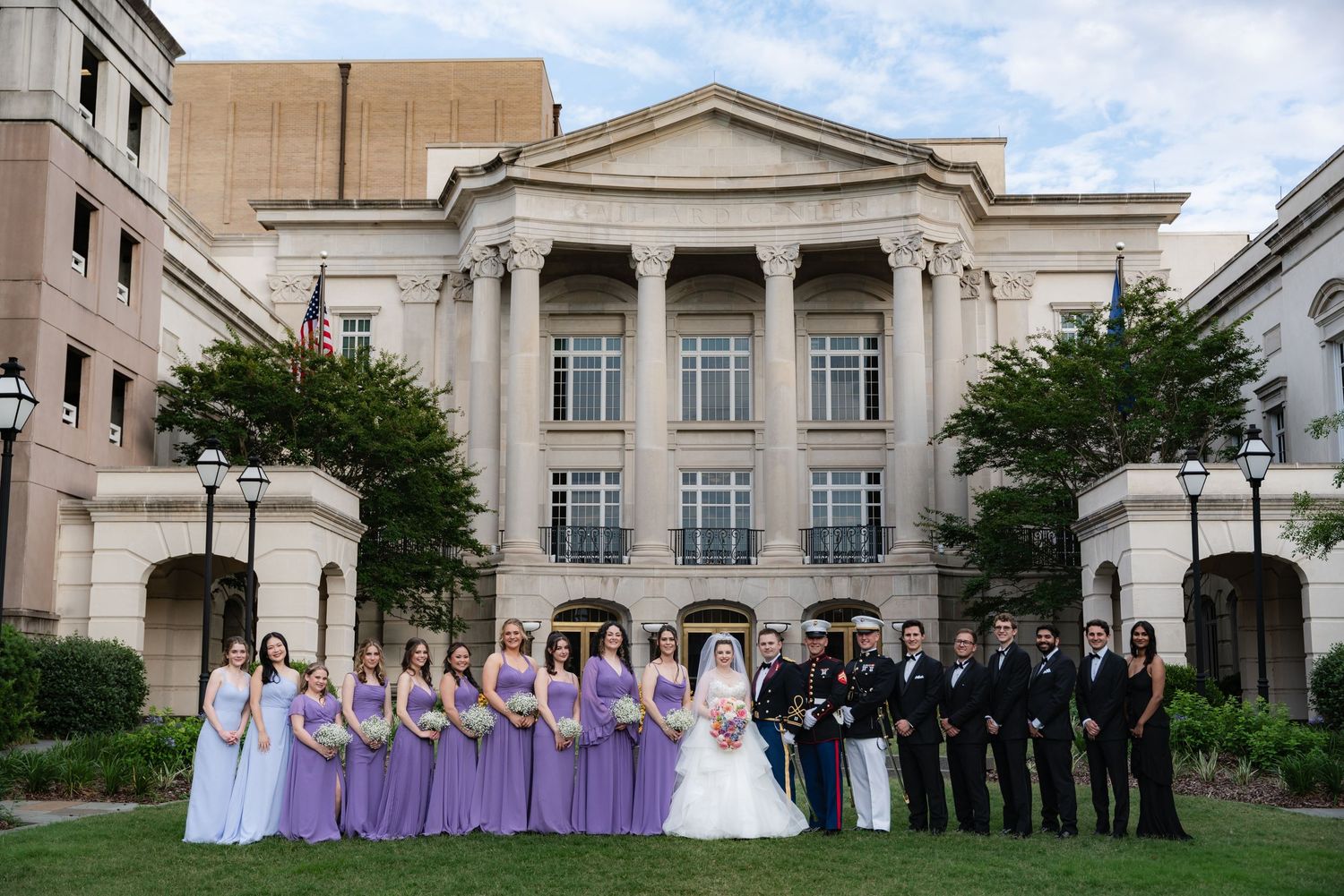 A wedding party poses together in formal attire outside a grand neoclassical building with white columns and symmetrical trees.