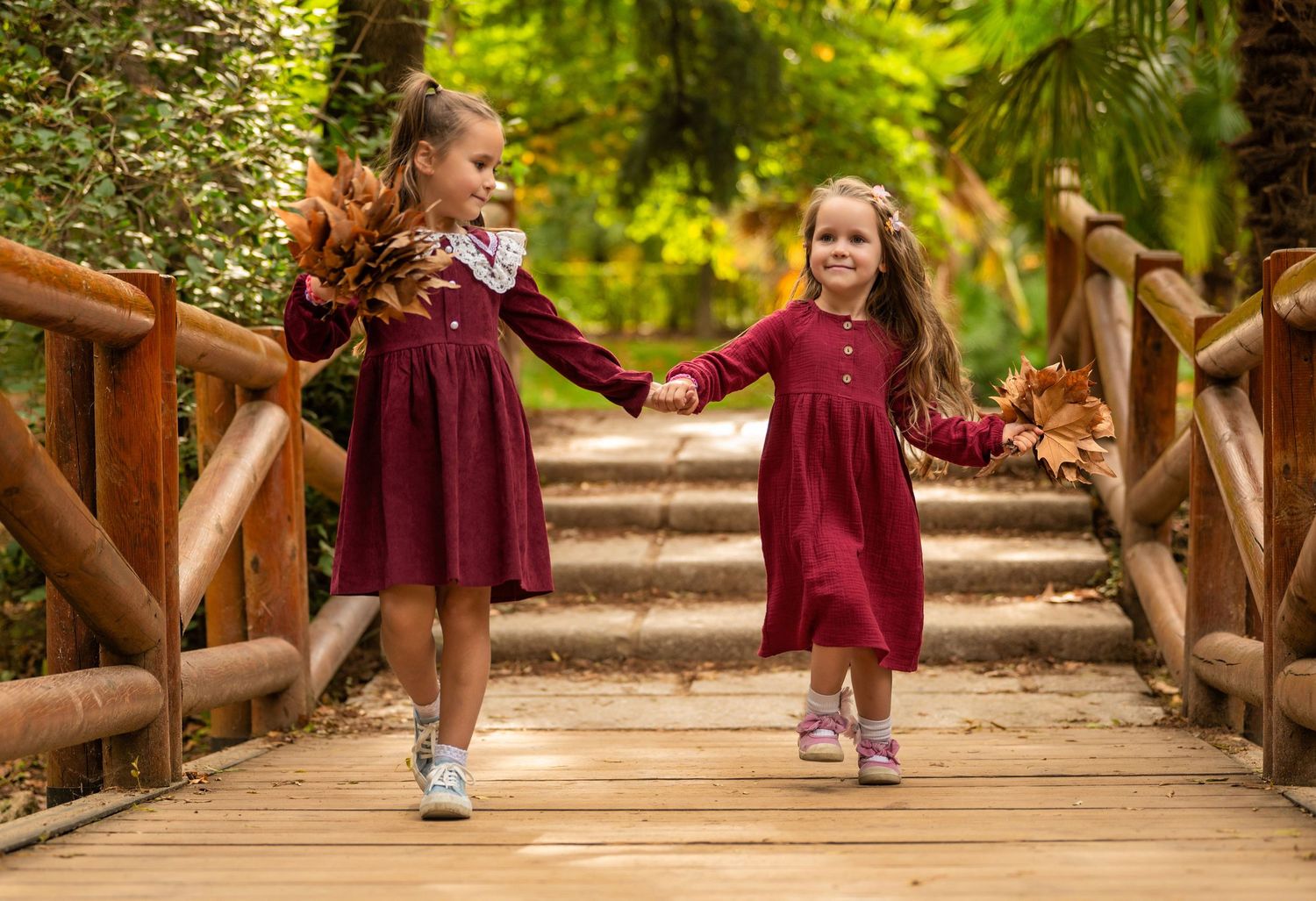 Two children in matching burgundy dresses hold hands while walking across a wooden footbridge in a lush garden setting.