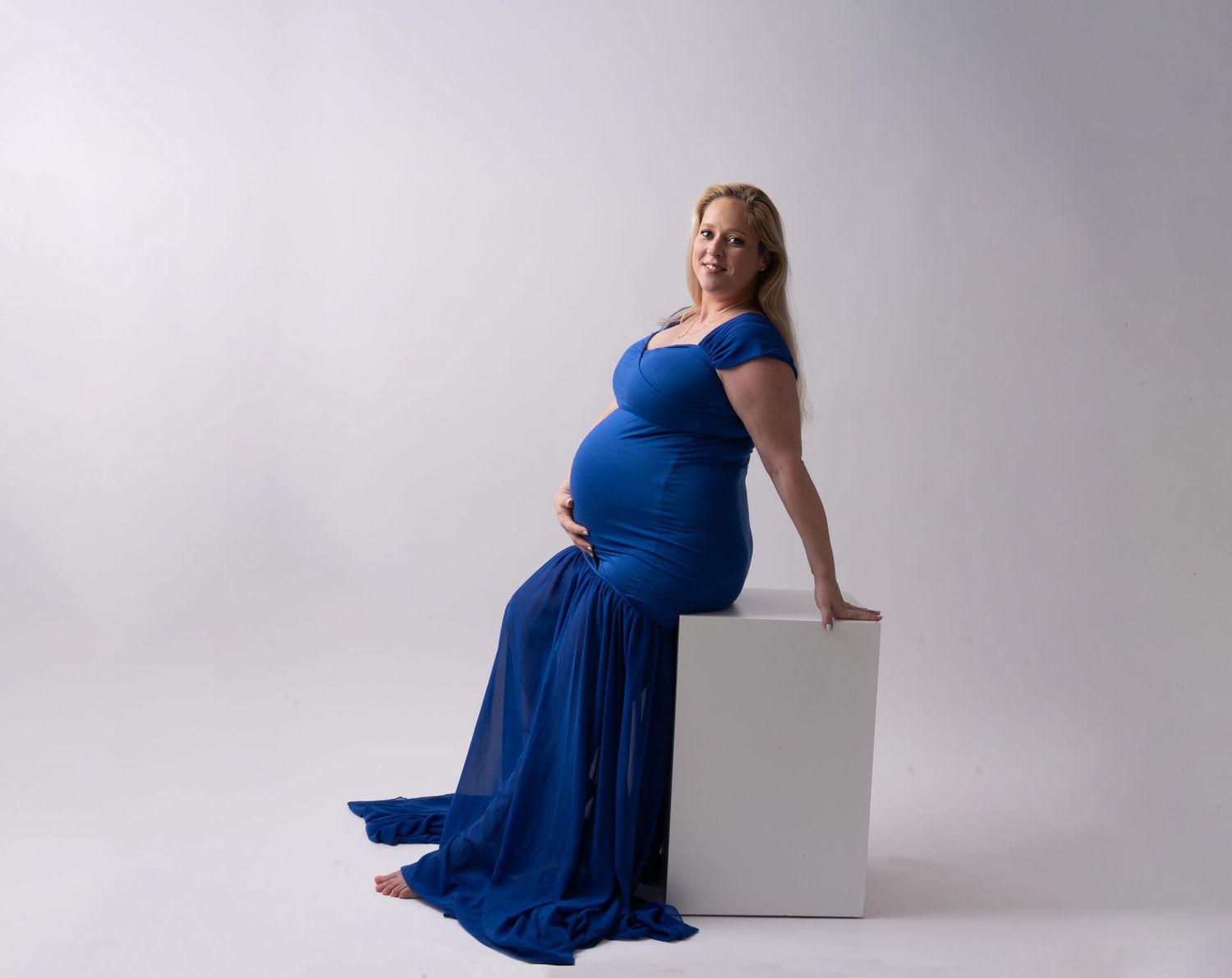 Expectant mother in flowing royal blue gown poses elegantly while seated on white cube during maternity photoshoot.