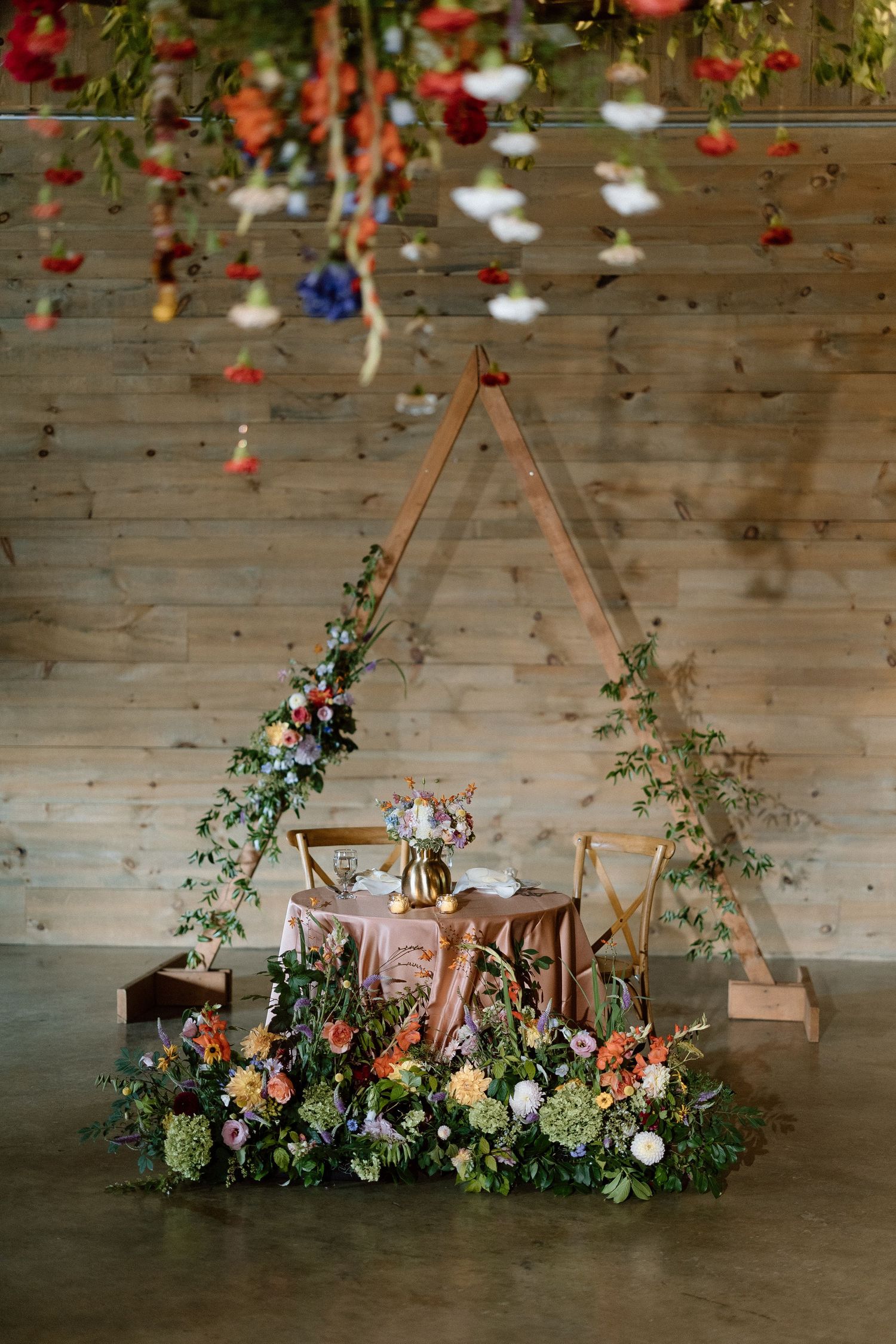 A boho-style wood triangle arch adorned with blush and white floral arrangements on a rustic wooden floor.