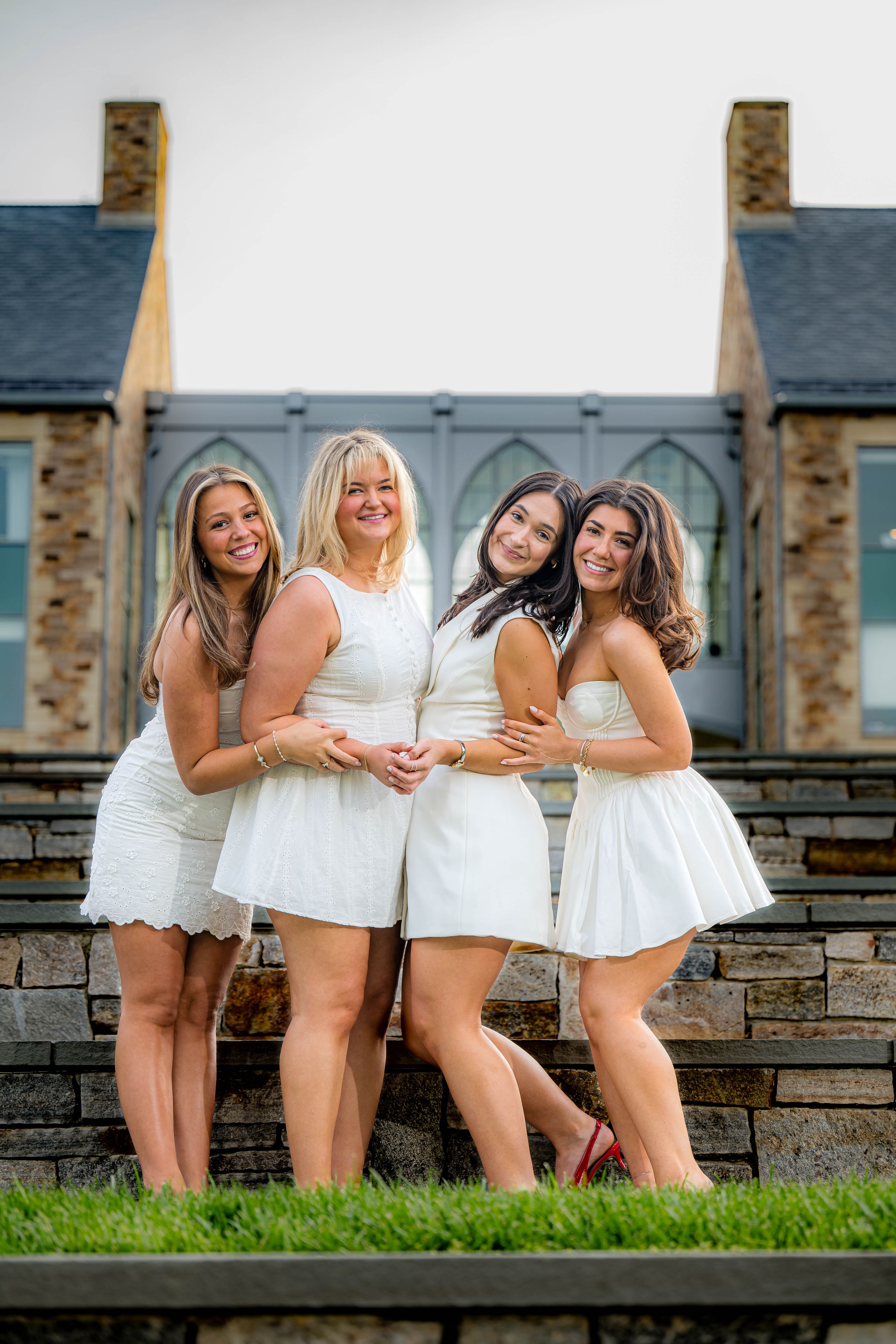 Four friends in matching white dresses pose together on stone stairs at an outdoor venue.