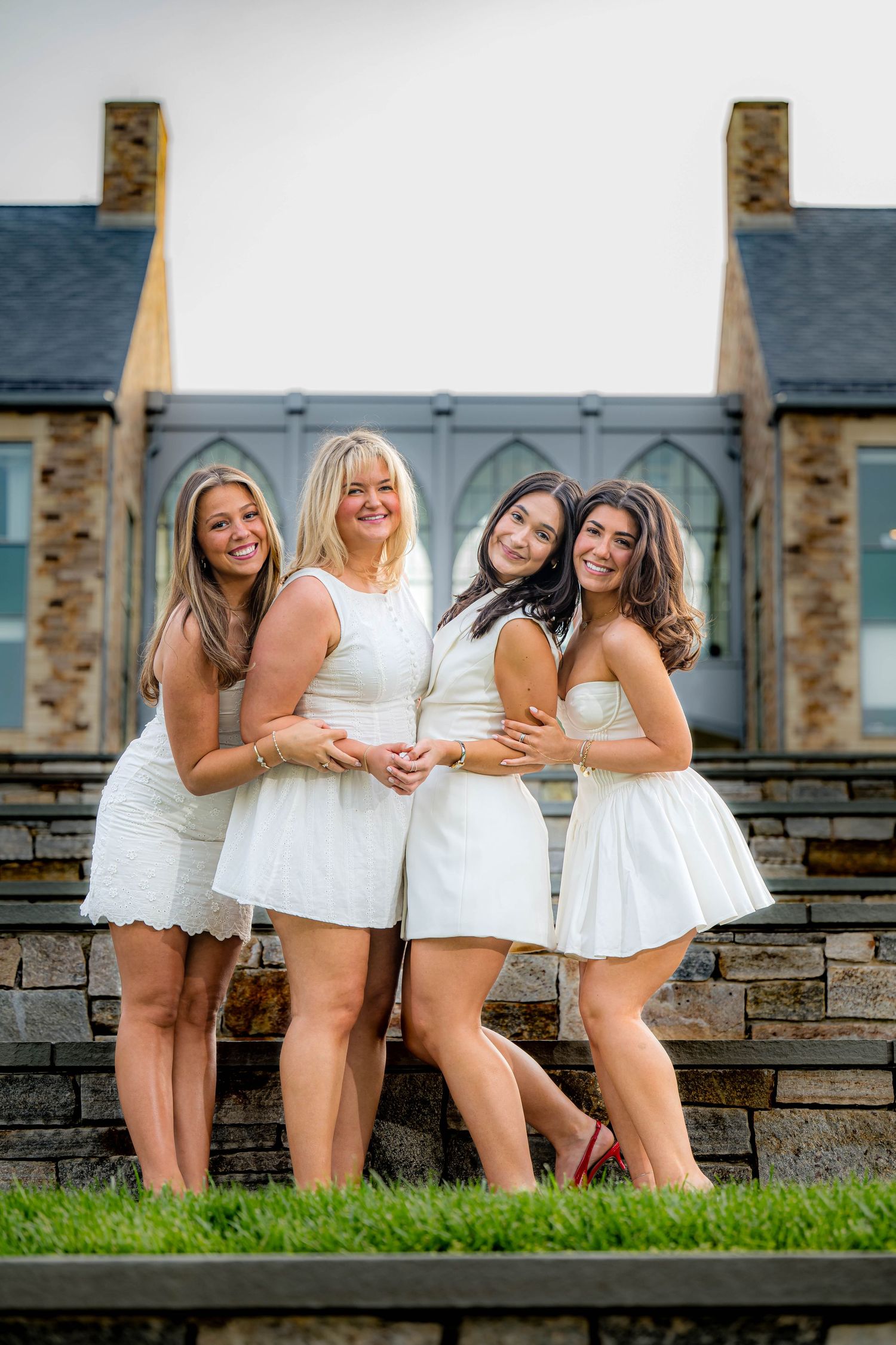 Four friends in matching white dresses pose together on stone stairs at an outdoor venue.
