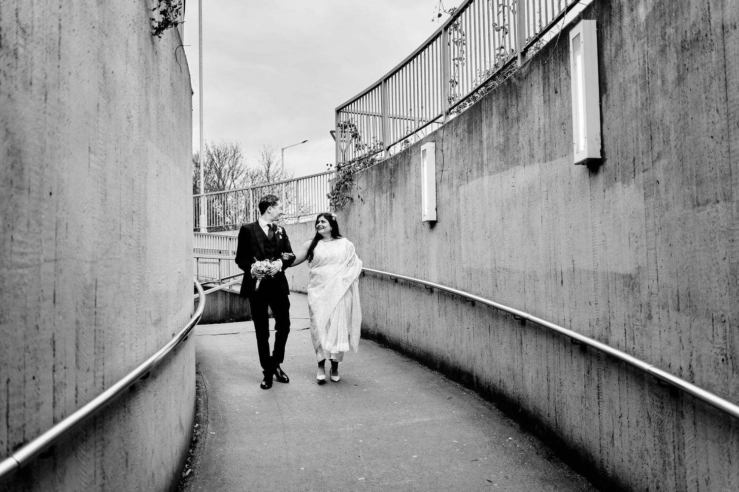Two people walking together down a concrete ramp between tall grey walls in black and white photography in Canterbury, Kent.