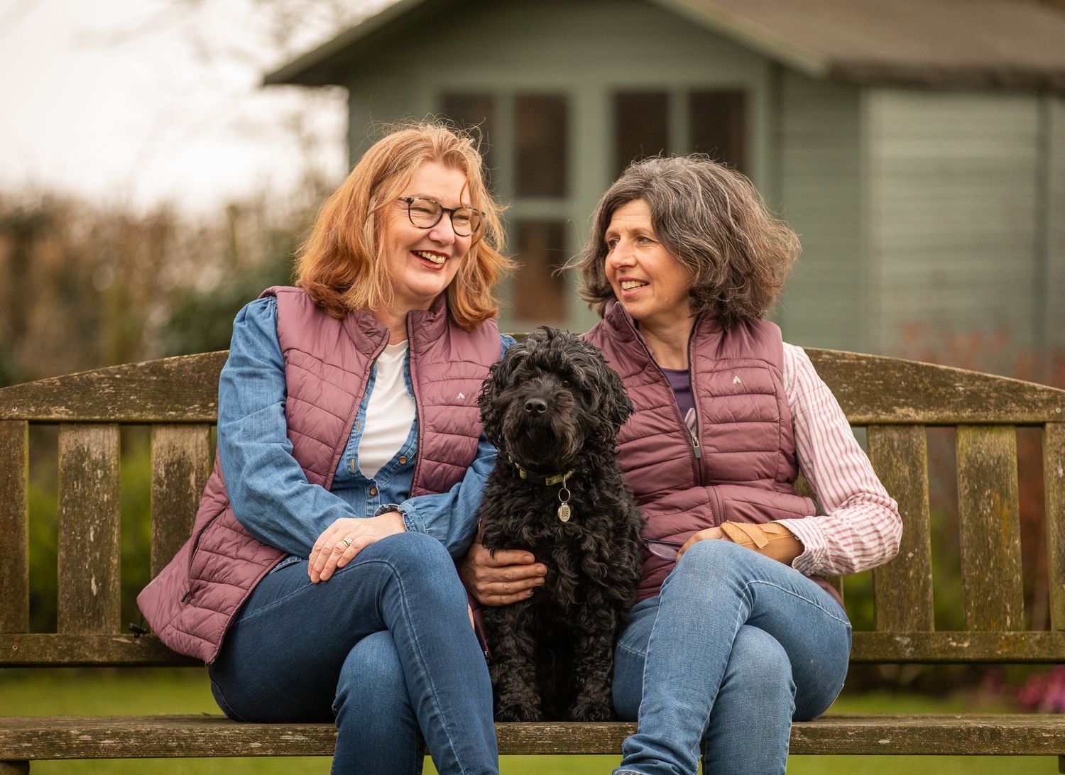 Two friends in puffer vests sit on a wooden bench with their black dog in a backyard garden.