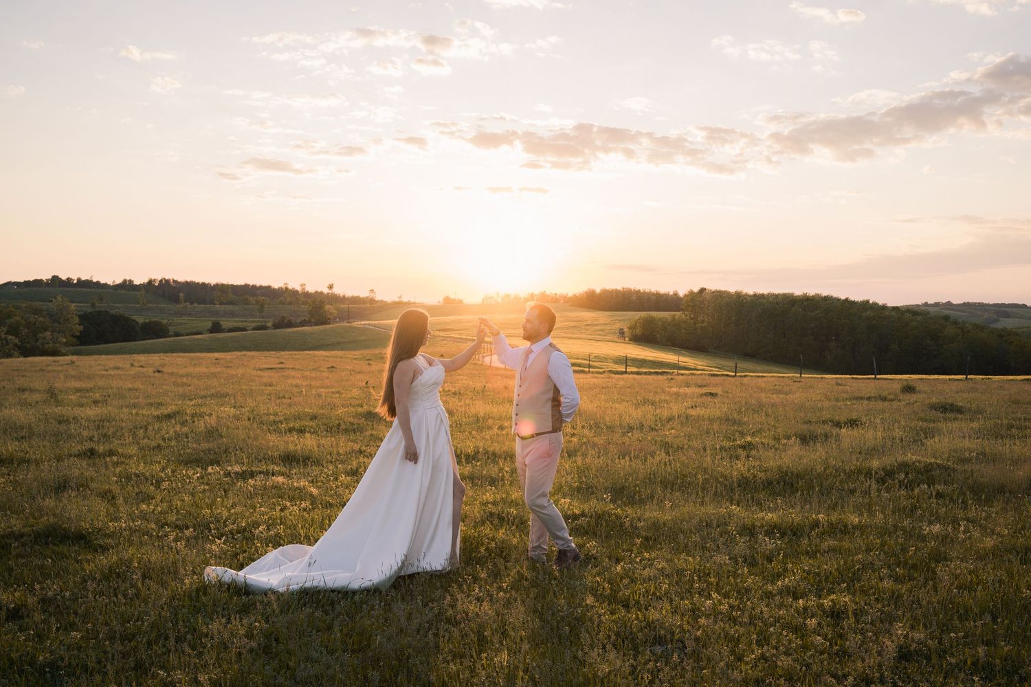 A couple in wedding attire embrace in a sun-drenched meadow at sunset with flowing white dress and golden light.