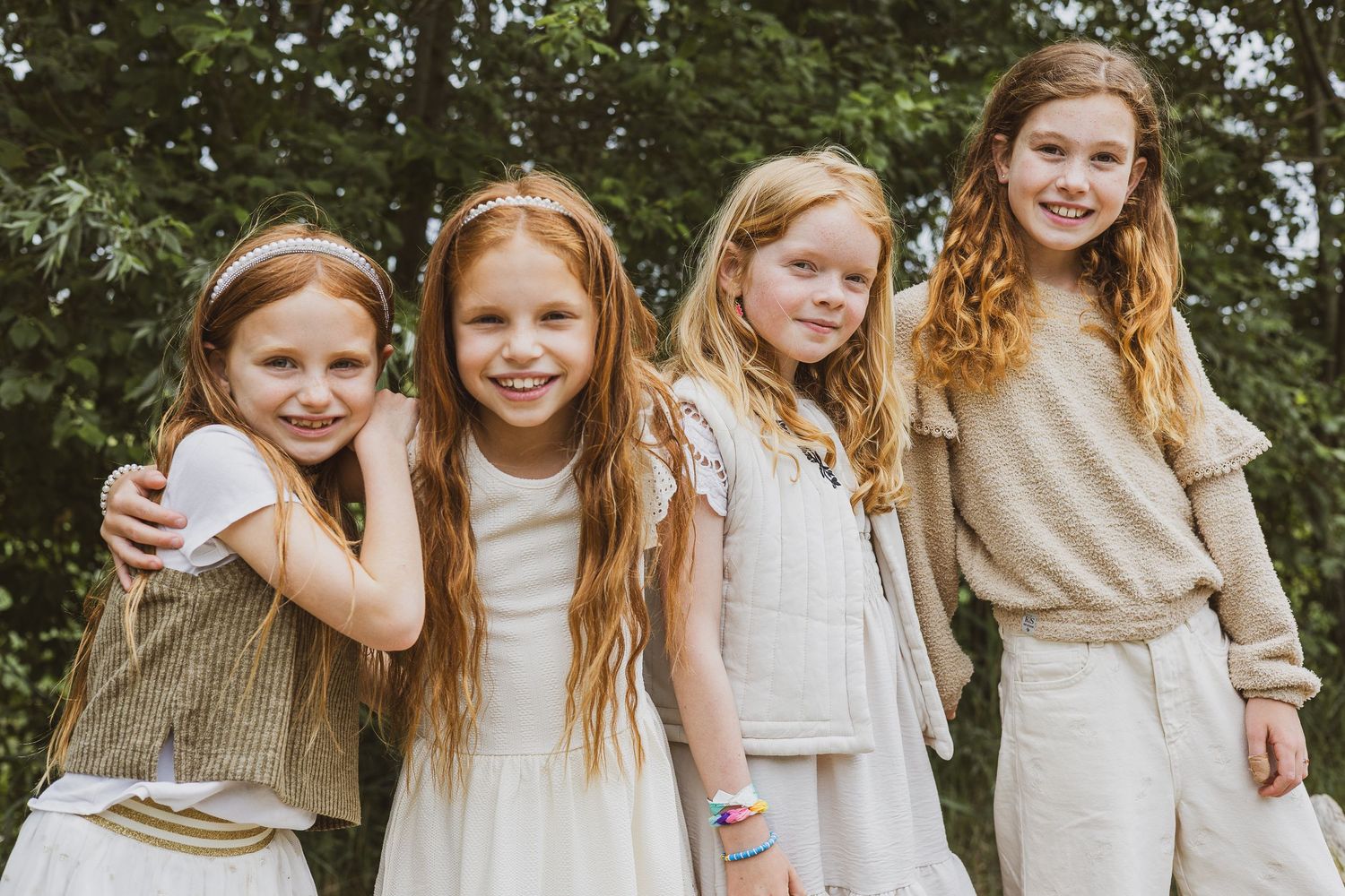 Four young friends standing together outdoors wearing neutral colored clothing in a natural setting.