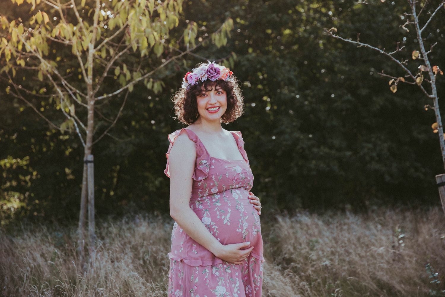 A pregnant woman in a pink floral dress stands in a sunlit field with tall grass in the background. During a maternity session in London