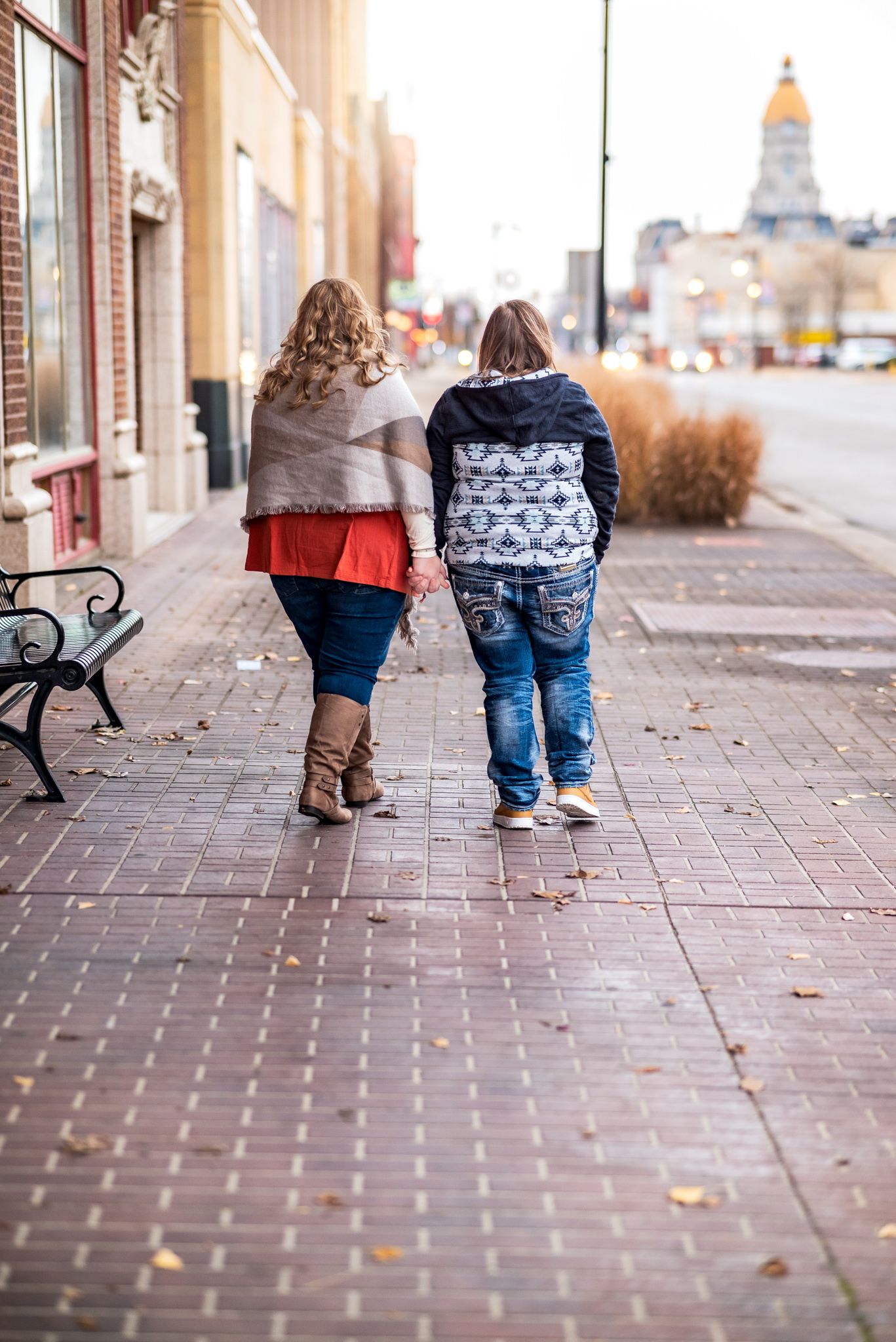 Two people walk together down a brick sidewalk on a small town main street with the state capitol dome visible in the distance.