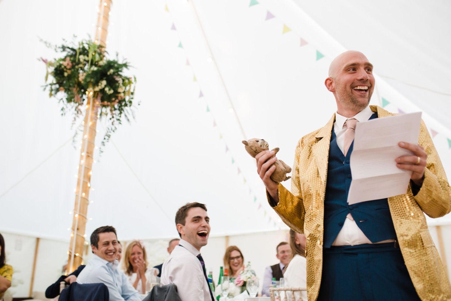 A bestman in a gold jacket holds papers and a teddy while addressing guests under a tent at Wilson Coastal Club NS wedding venue.