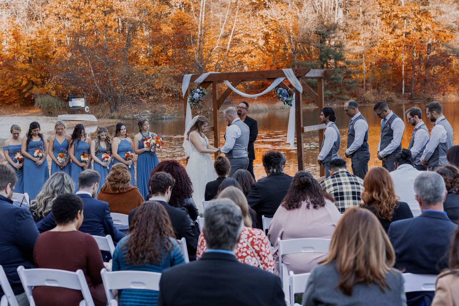 Outdoor wedding ceremony by a lake with vibrant autumn foliage and wooden arch during fall season.