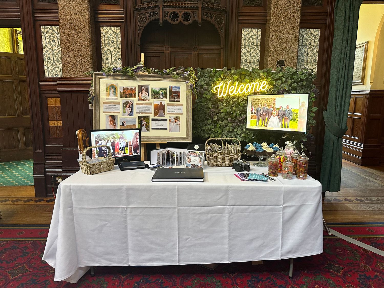 Wedding welcome table with photo display, neon sign and memorabilia against wood-paneled wall.