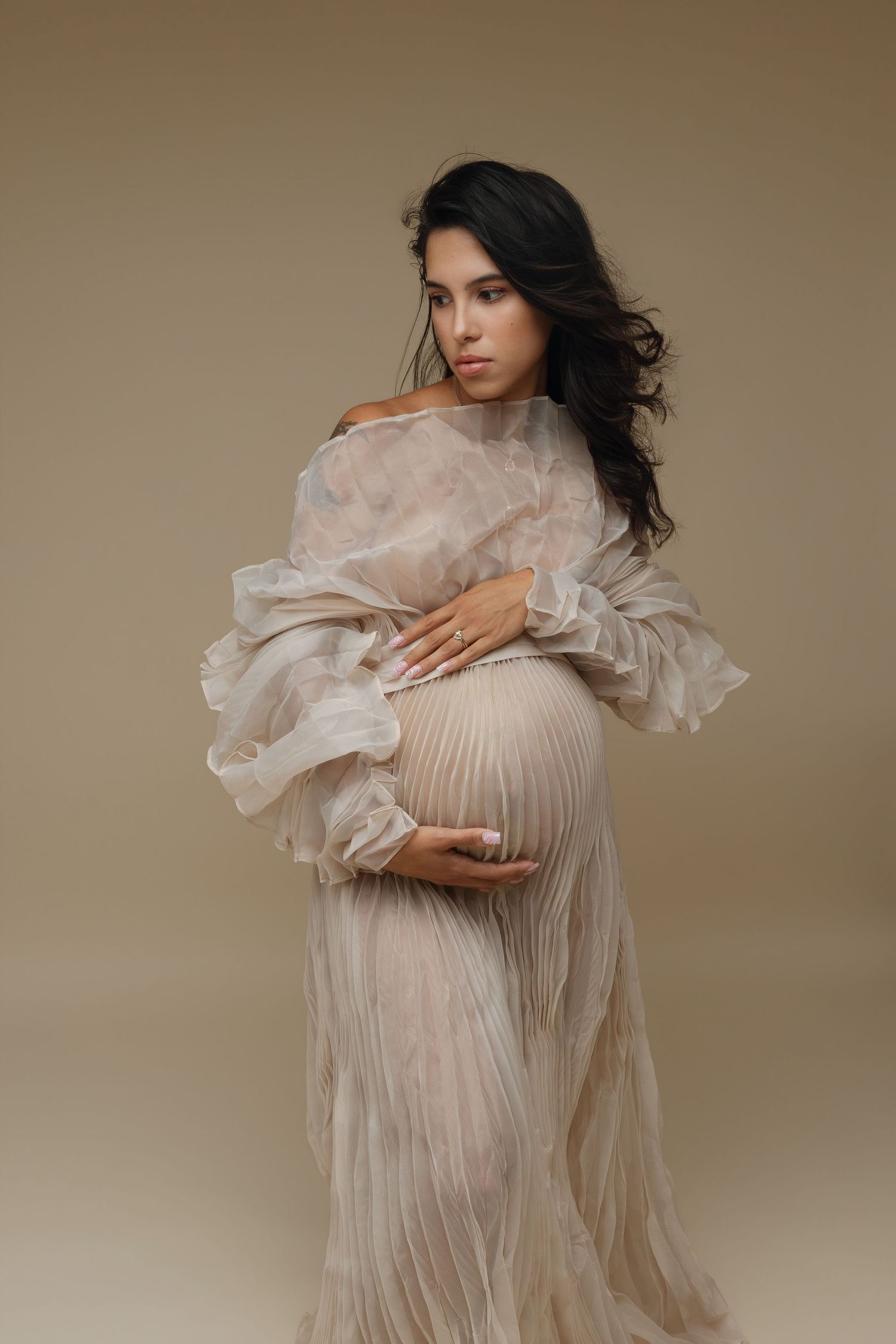 Pregnant woman in sheer beige pleated gown cradling baby bump, elegant maternity portrait on neutral background.