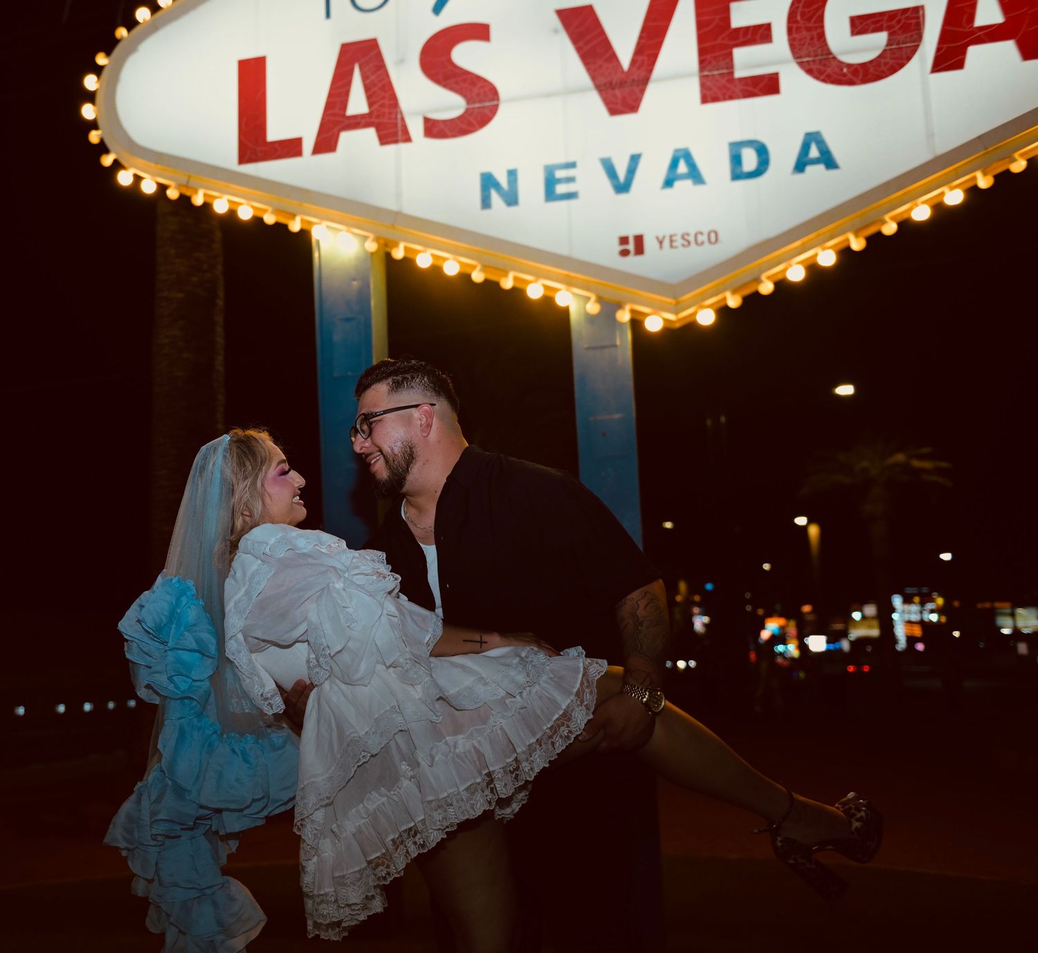 Couple celebrating wedding at iconic Welcome to Las Vegas Nevada sign at night, groom lifting bride in white dress.