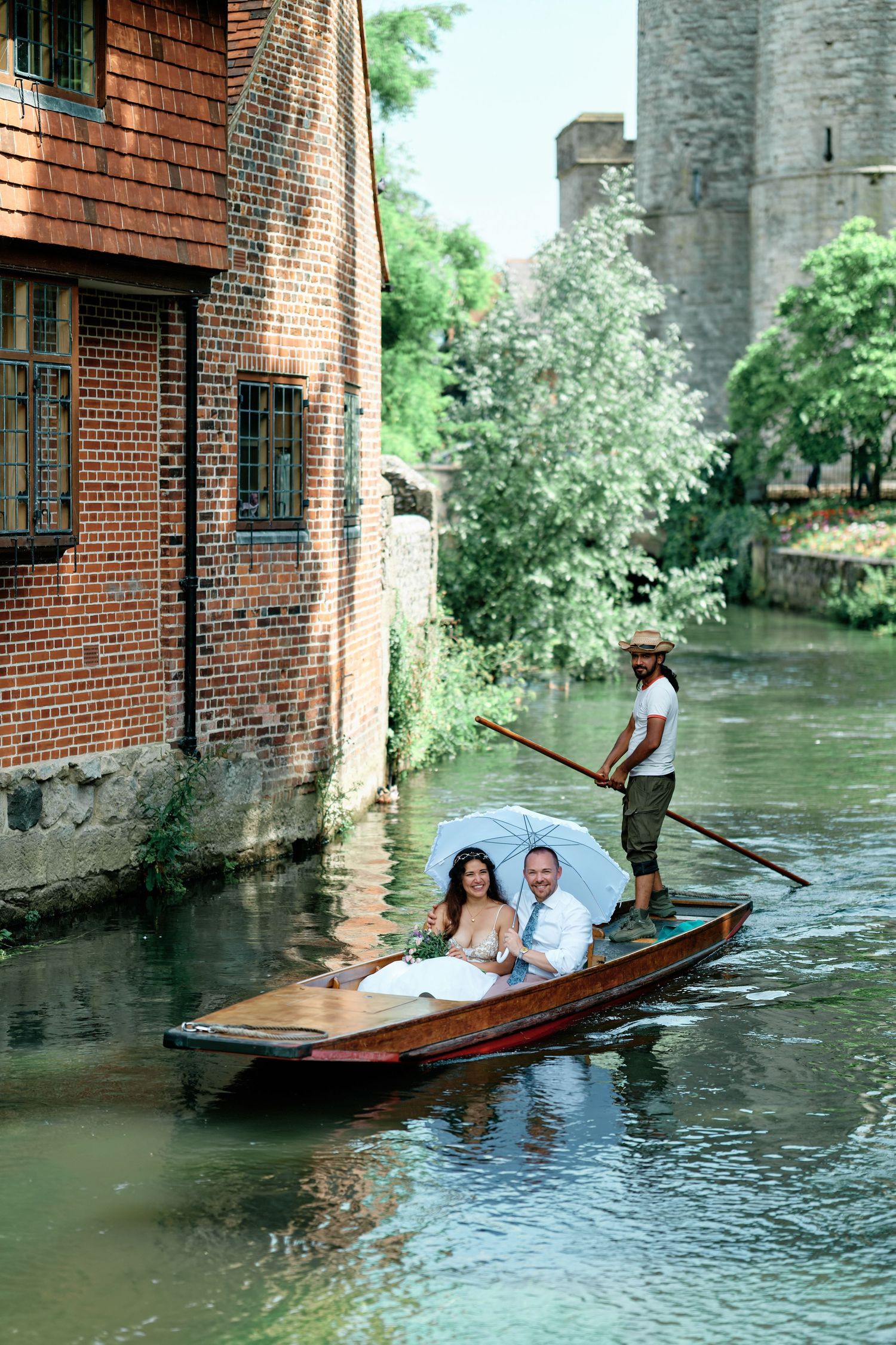 Couple in wedding attire riding a punt on a scenic canal, guided by a gondolier past brick buildings in Canterury