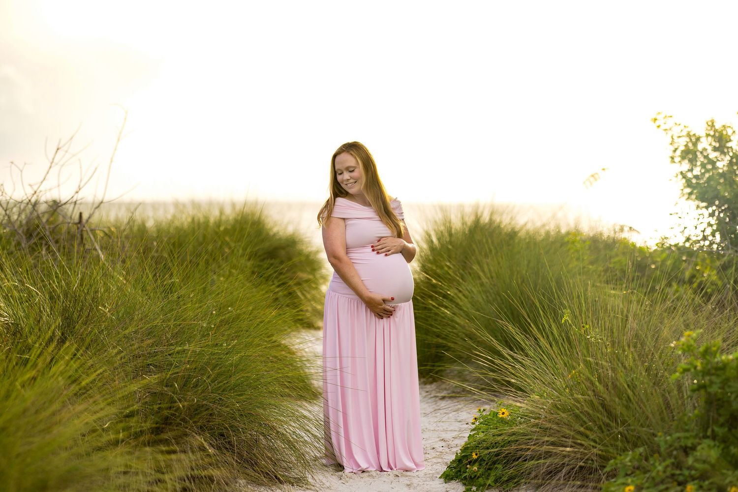 Pregnant woman in pink maxi dress smiling on sandy beach path surrounded by tall green grass at sunset.