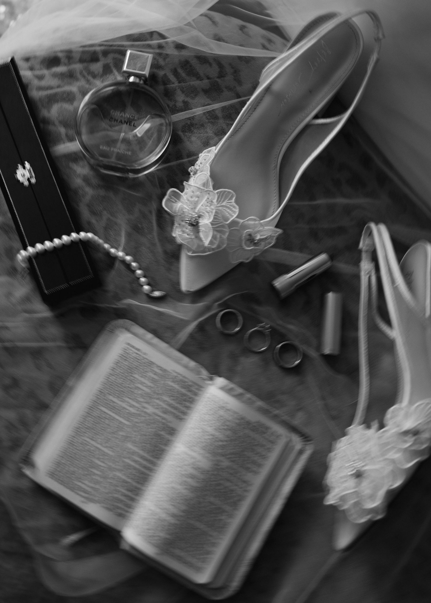 Black and white wedding flat lay with bridal heels, pearl necklace, rings, perfume, lipstick, and open Bible.