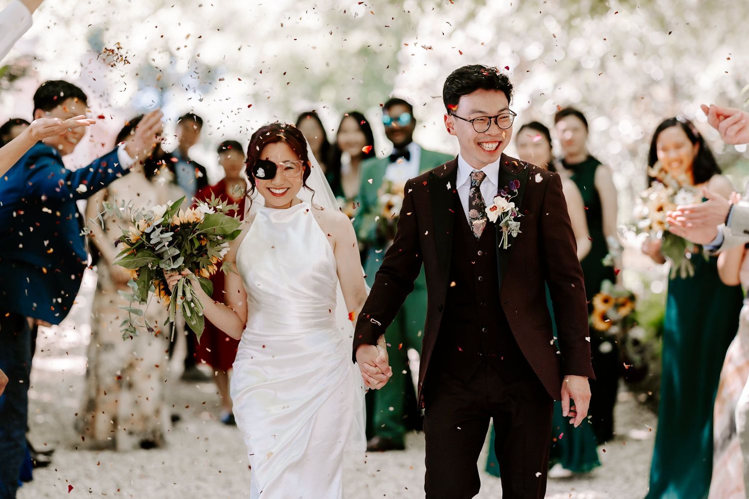 Asian couple walks through confetti shower after wedding ceremony, bride in white gown holding bouquet, groom in dark suit.