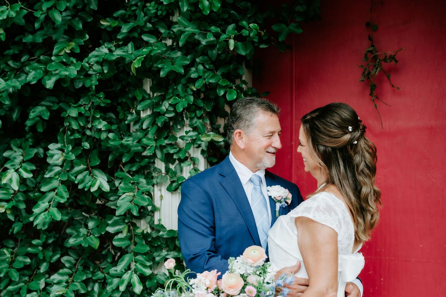 A bride and groom share a romantic moment in front of a red barn wall covered in lush green ivy.