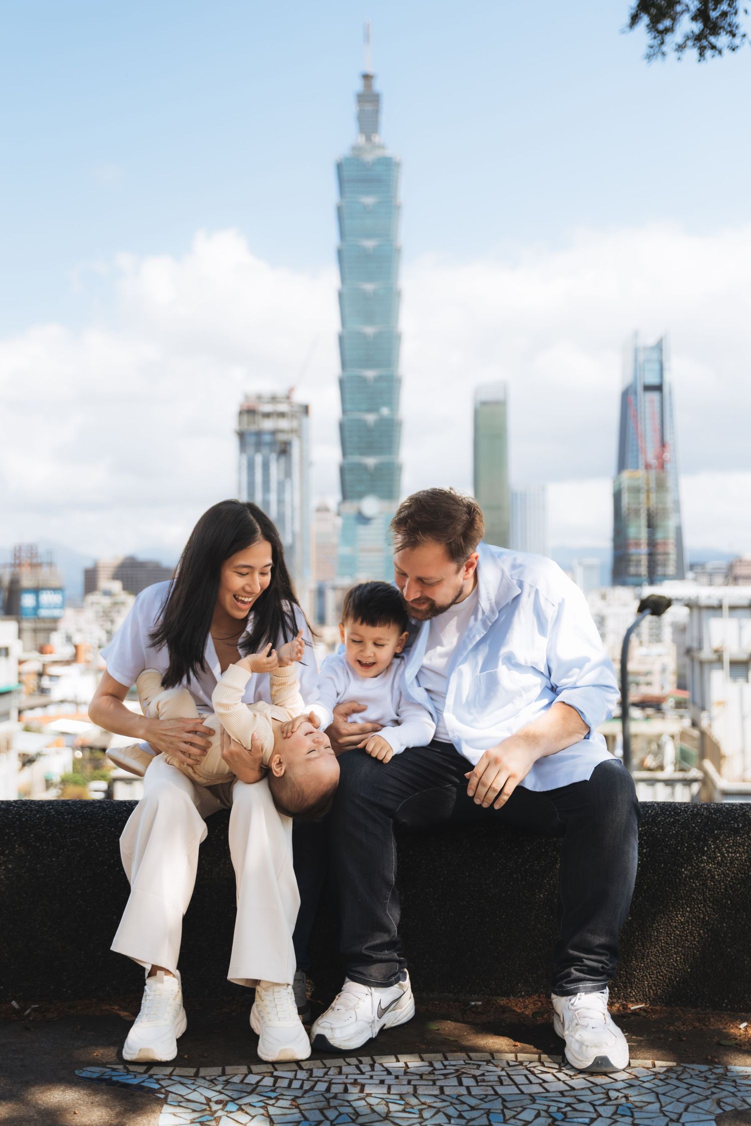 Happy family of four posing with Taipei 101 skyline backdrop, raising hands joyfully on a sunny day.